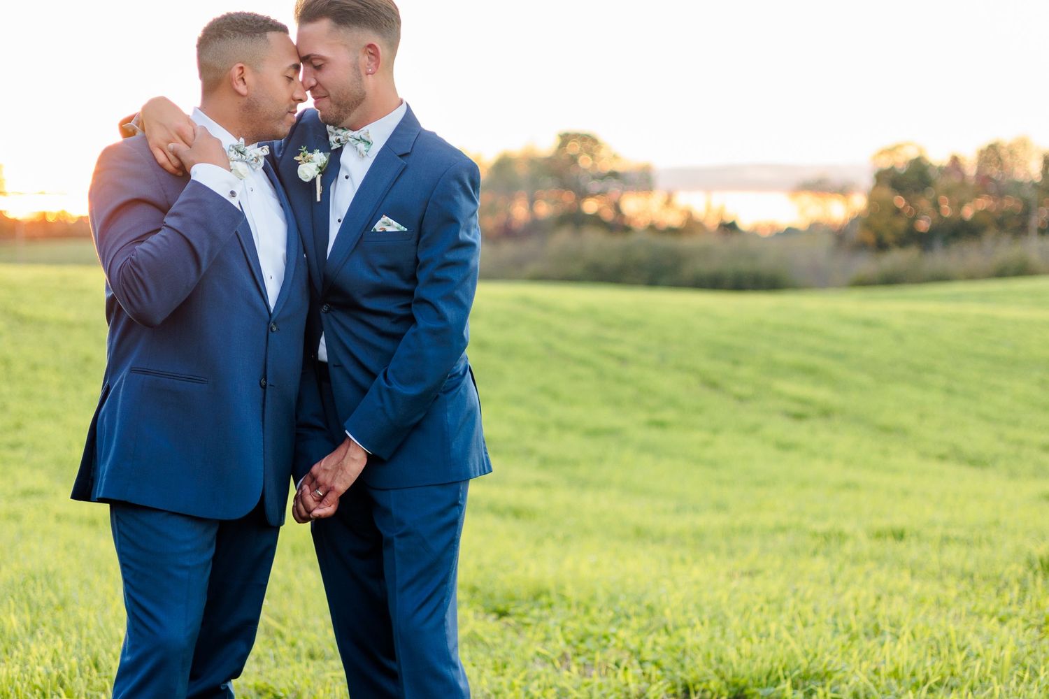 Couple in blue suits sharing intimate moment in grassy field at sunset.
