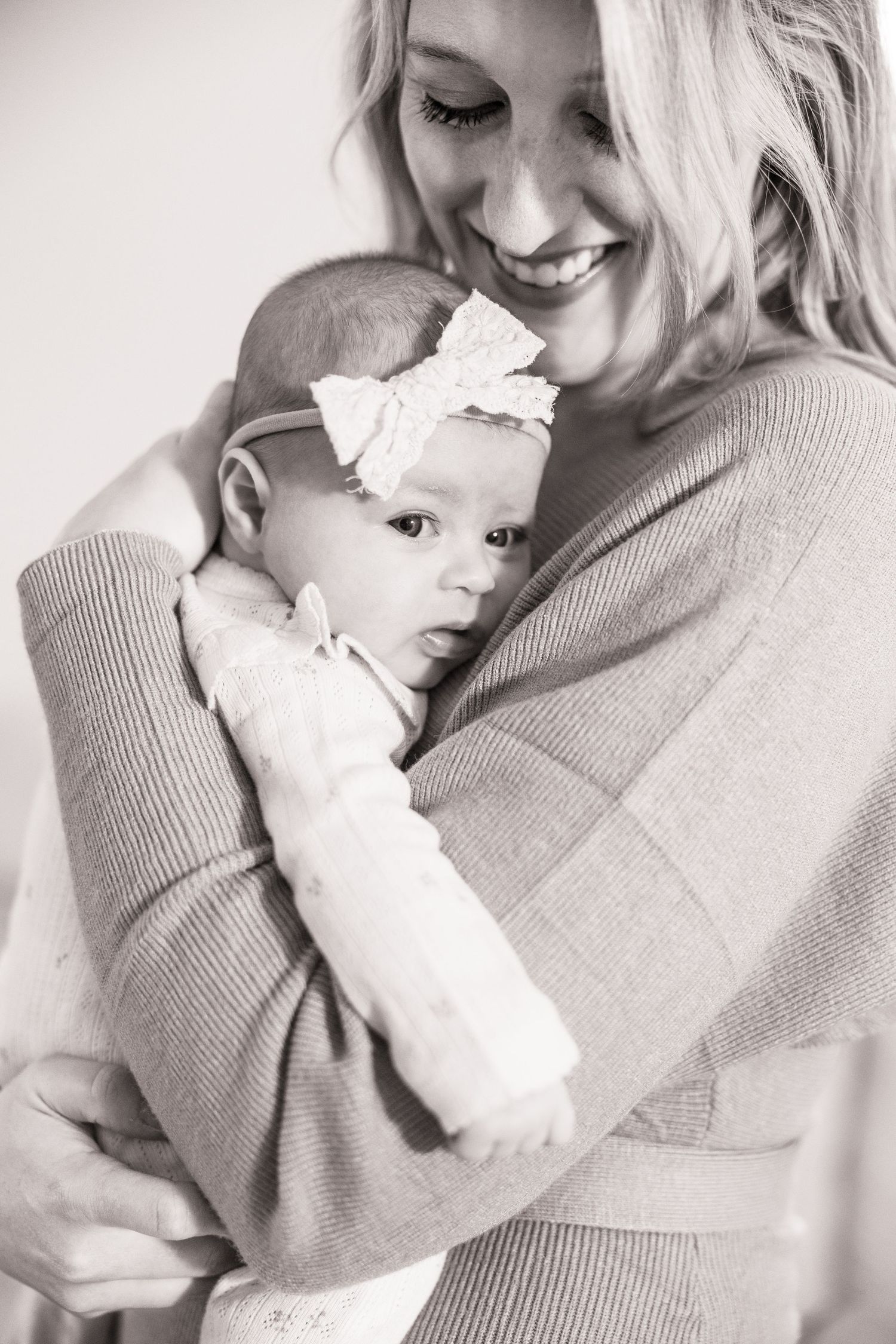Black and white portrait of a mother in a sweater embracing her baby wearing a white bow headband.