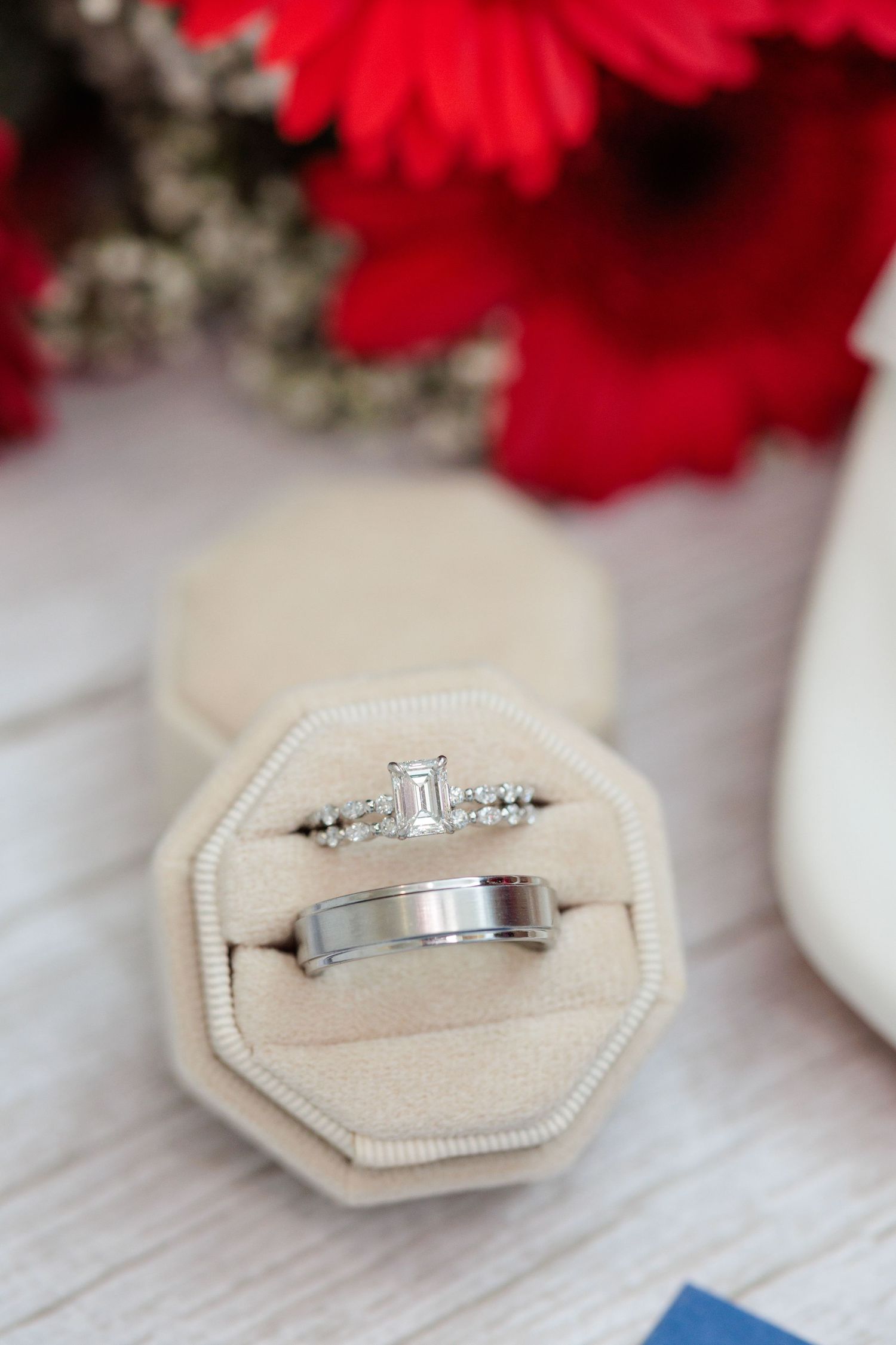 Elegant diamond engagement ring displayed on beige cushion with red flowers in background.