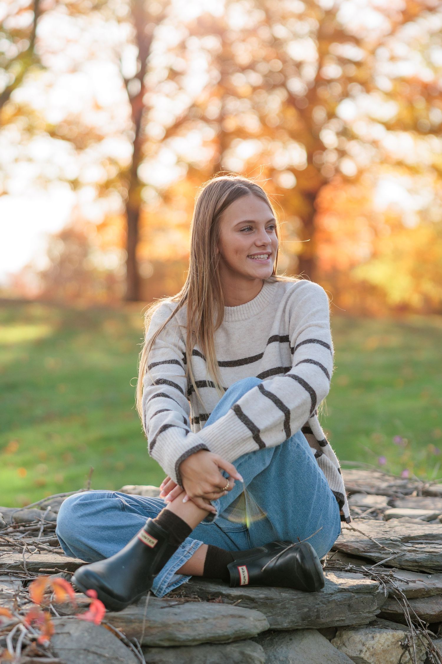 Young woman in striped sweater and jeans sitting on rocks outdoors during autumn with colorful fall foliage background.