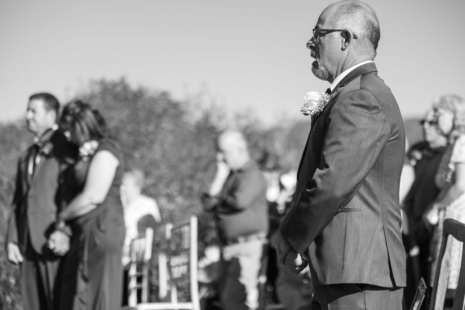 Black and white photo of suited man standing solemnly among seated wedding guests during outdoor ceremony.