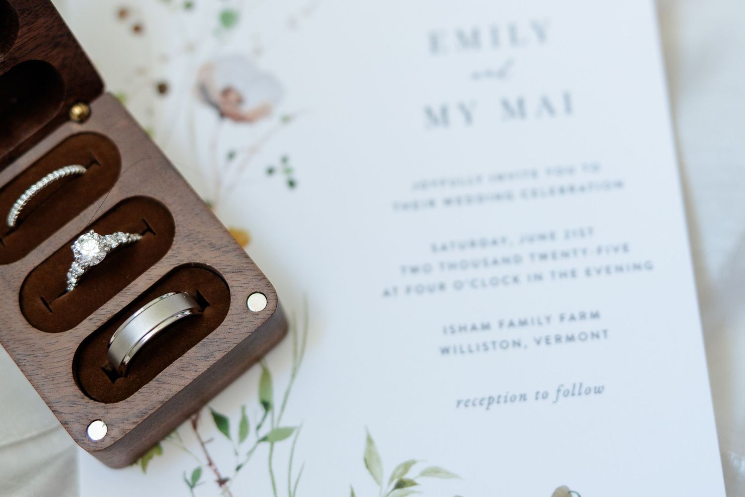 Wedding rings in a walnut wooden box beside an elegant floral wedding invitation for Emily and My Mai.