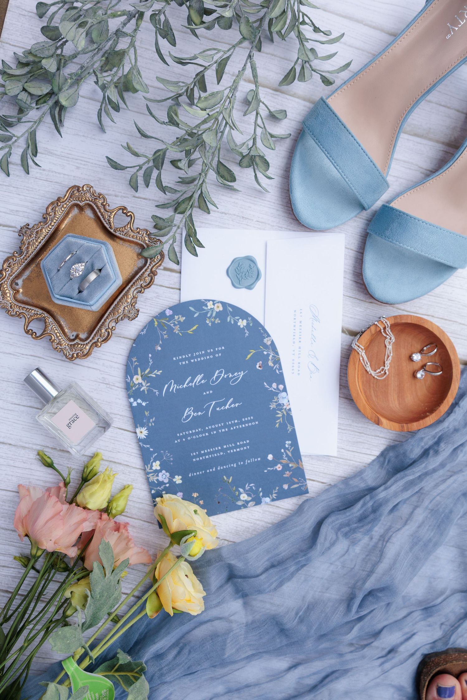 Blue wedding invitation flatlay with heels, roses, rings, perfume, and eucalyptus on white wood.