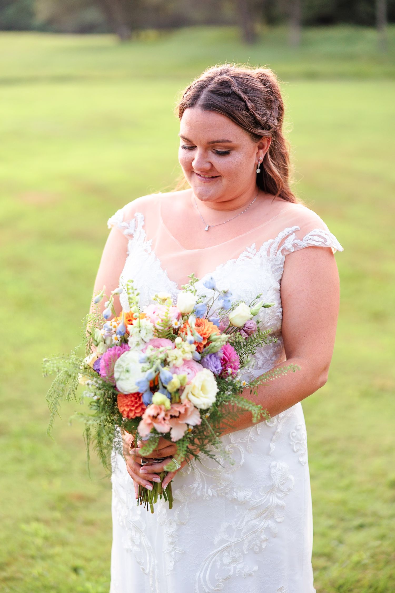 Smiling bride in lace wedding gown holding a colorful wildflower bouquet on a green lawn.