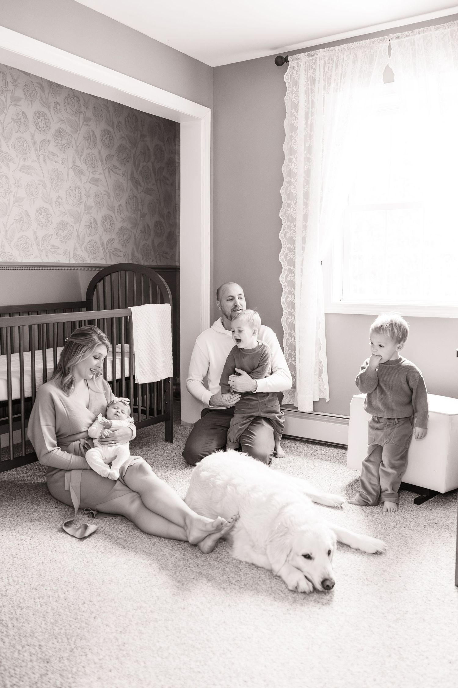 Family with two boys, newborn baby, and golden retriever dog relaxing together in nursery, black and white.