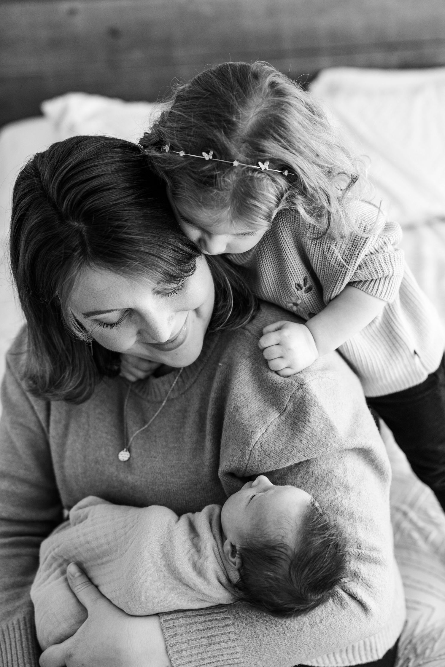 Black and white photo of mother holding newborn while toddler daughter kisses her cheek.