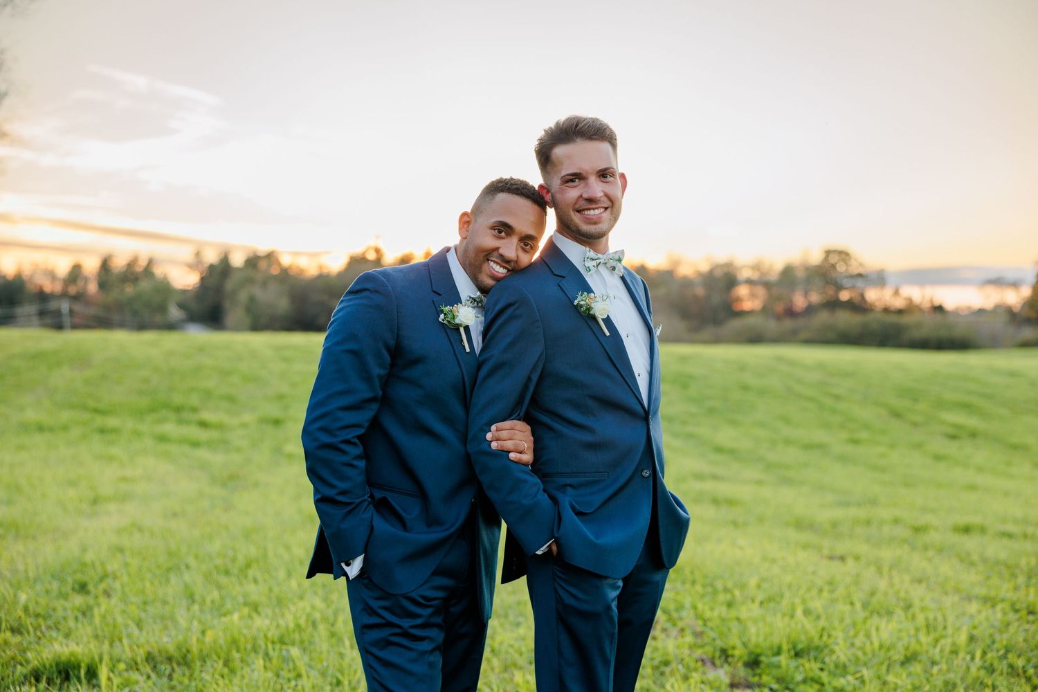 Two people in navy suits embrace during an outdoor wedding ceremony at sunset.