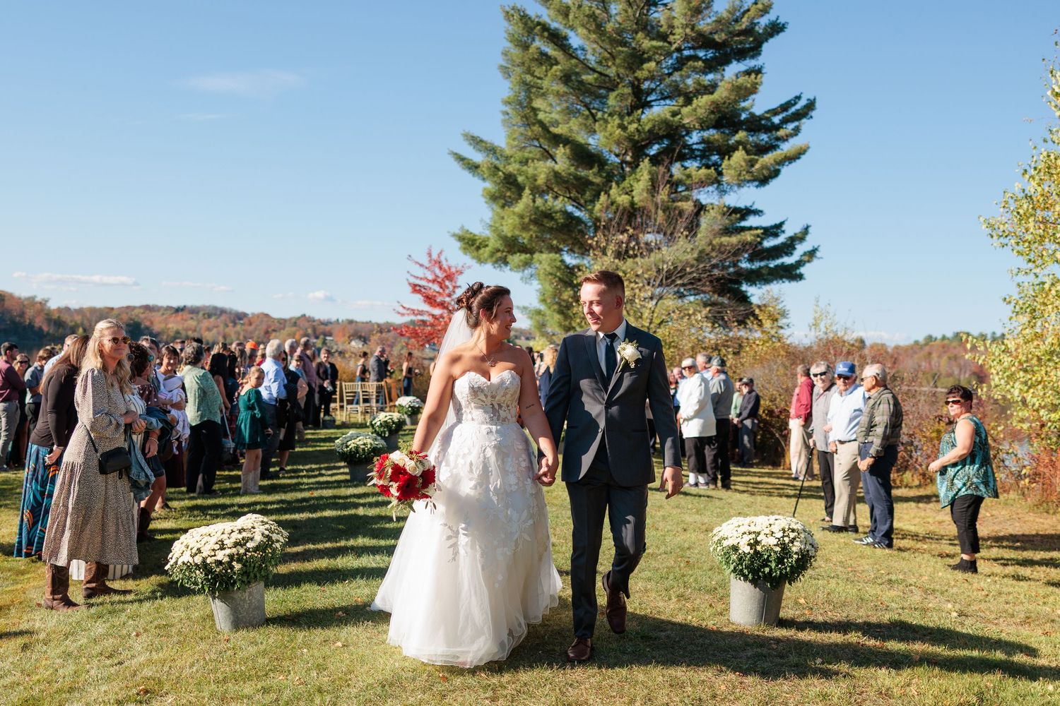 Wedding guests gather outdoors under blue skies with pine trees and fall foliage in the background.