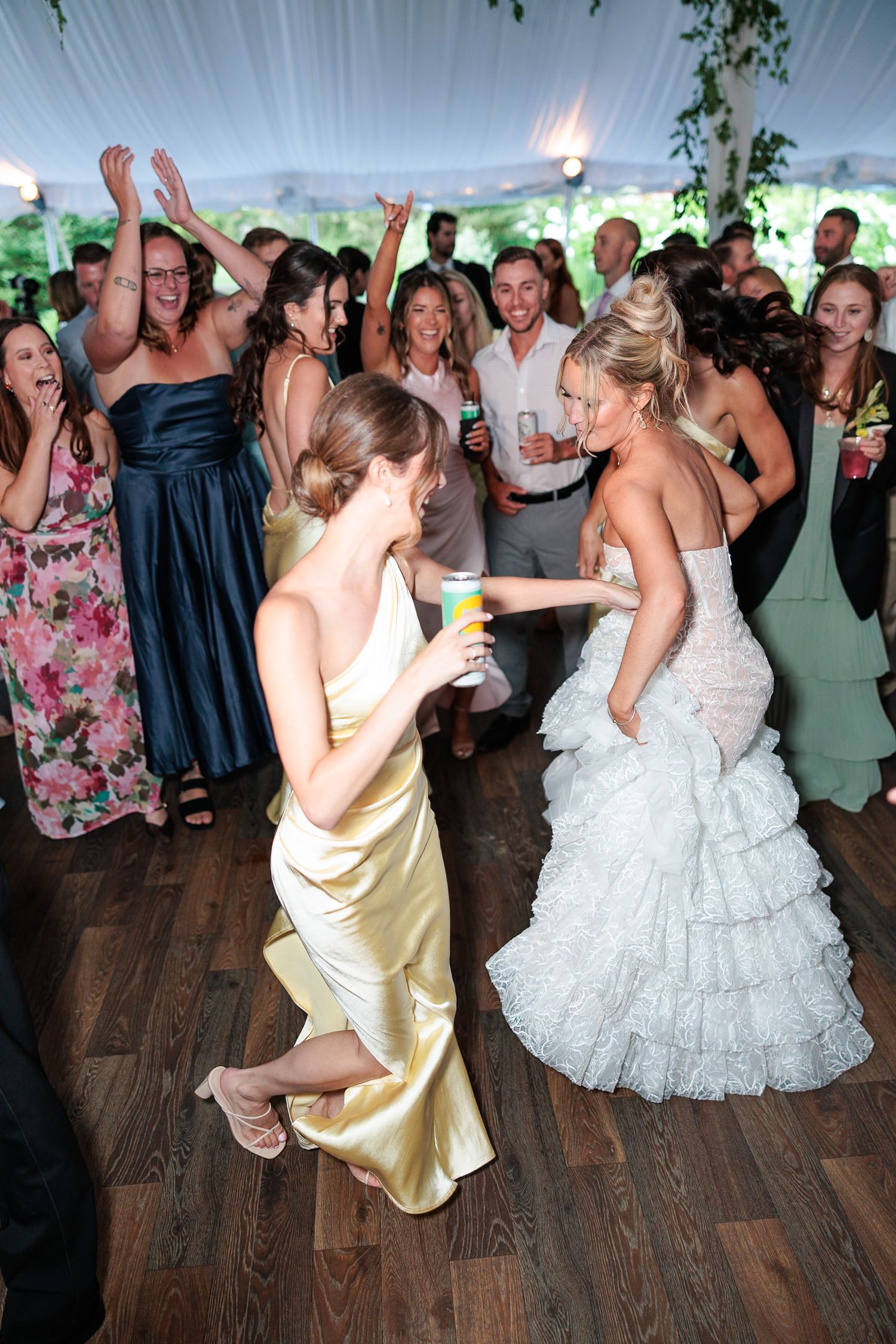 Bridesmaid in gold dress kneeling while dancing with bride in ruffled white gown at reception.