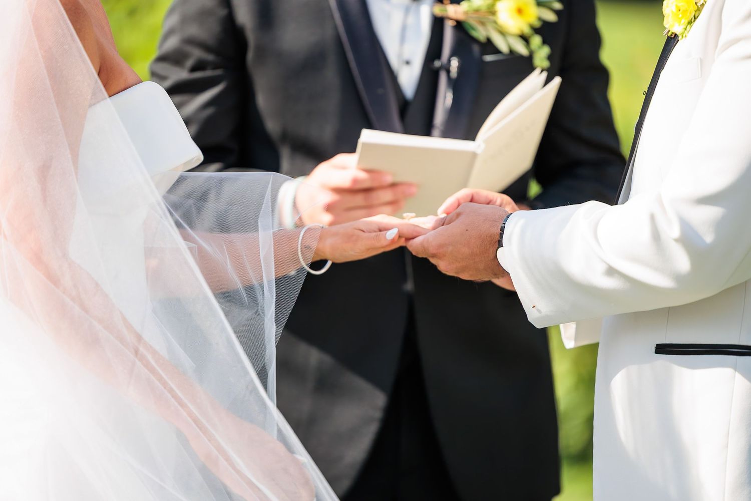 Close-up of groom placing wedding ring on brides finger during outdoor ceremony with officiant.