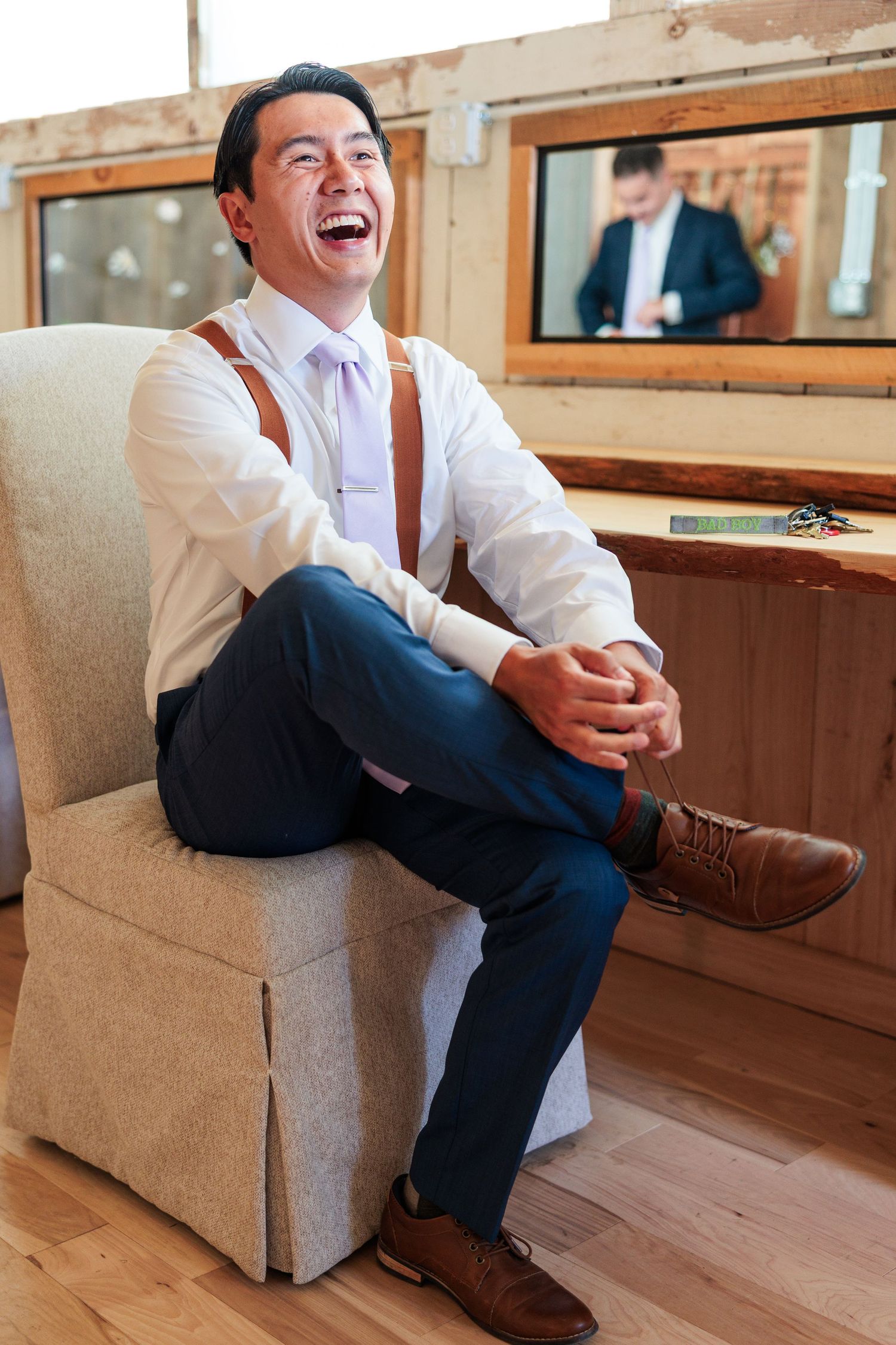 Laughing groom in white shirt and suspenders sitting in armchair during wedding preparations.