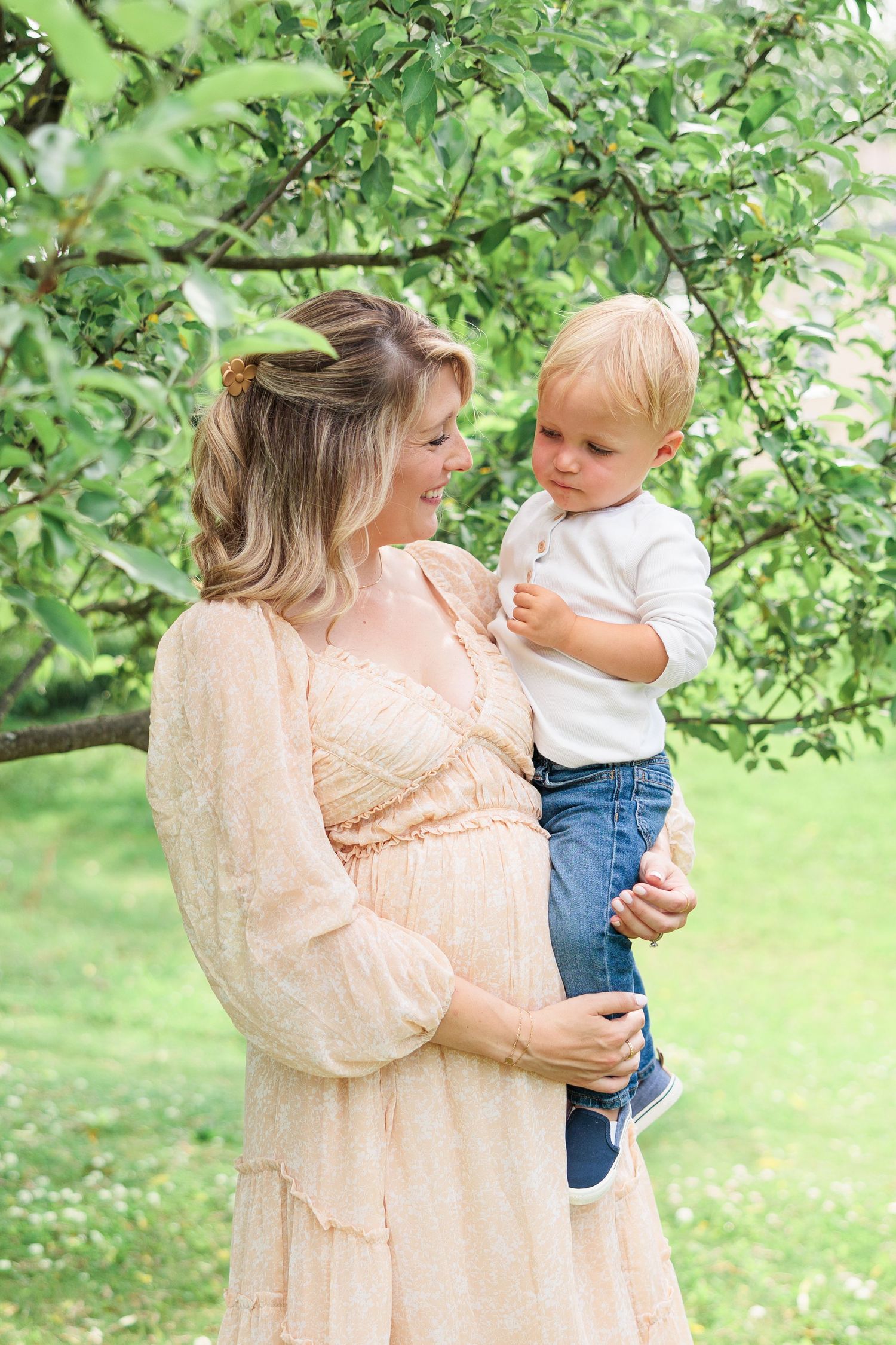 Pregnant woman in peach lace dress holding toddler son under green apple tree outdoors.
