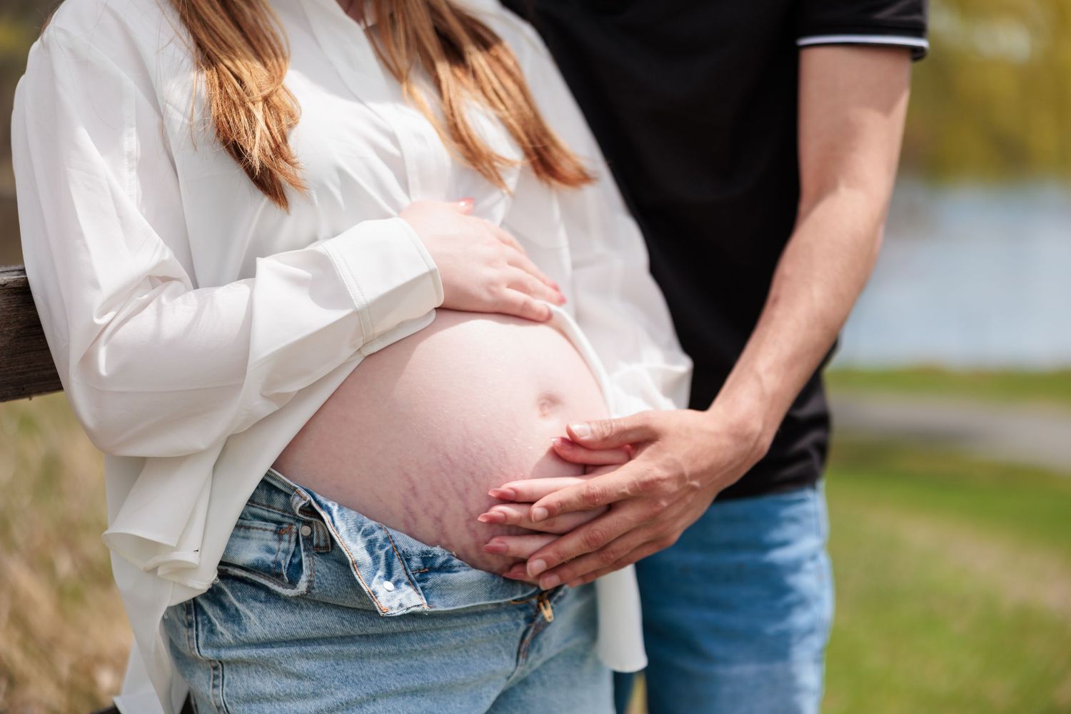 Close-up of couple's hands cradling pregnant woman's bare baby bump outdoors near a pond.