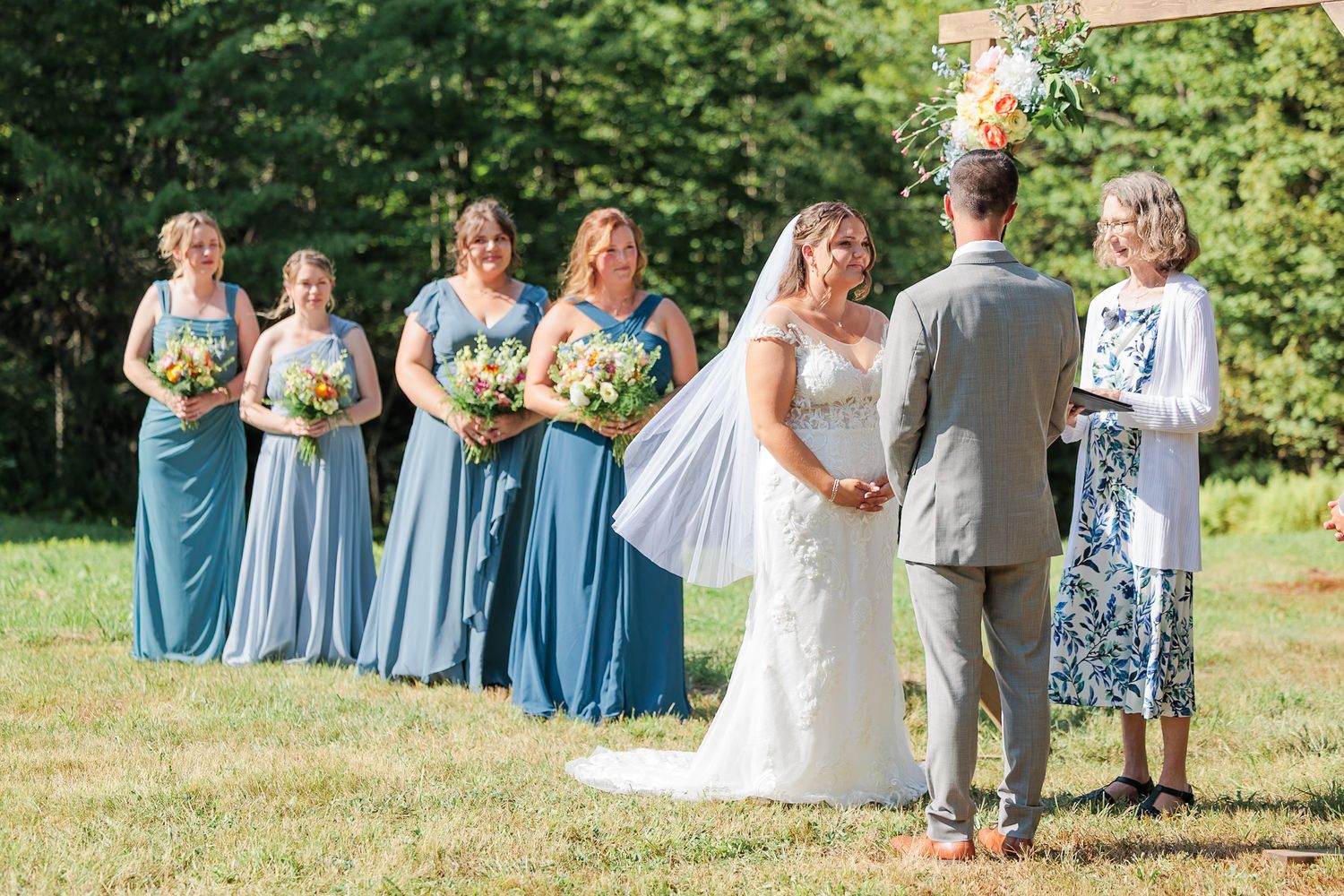 Bride exchanges vows with groom as bridesmaids in blue dresses stand beside her at outdoor wedding ceremony.