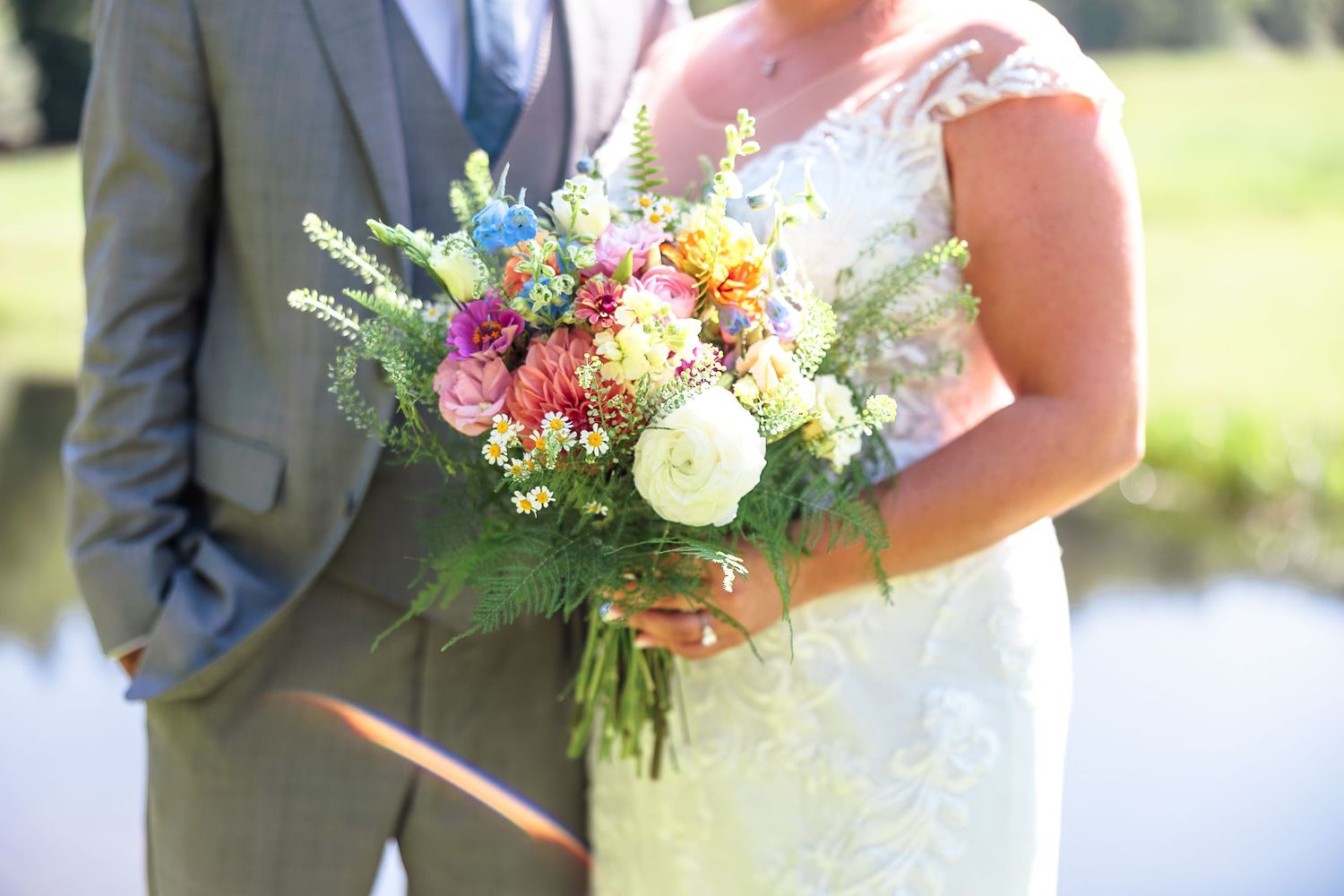 Bride and groom hold colorful wildflower wedding bouquet together near pond.