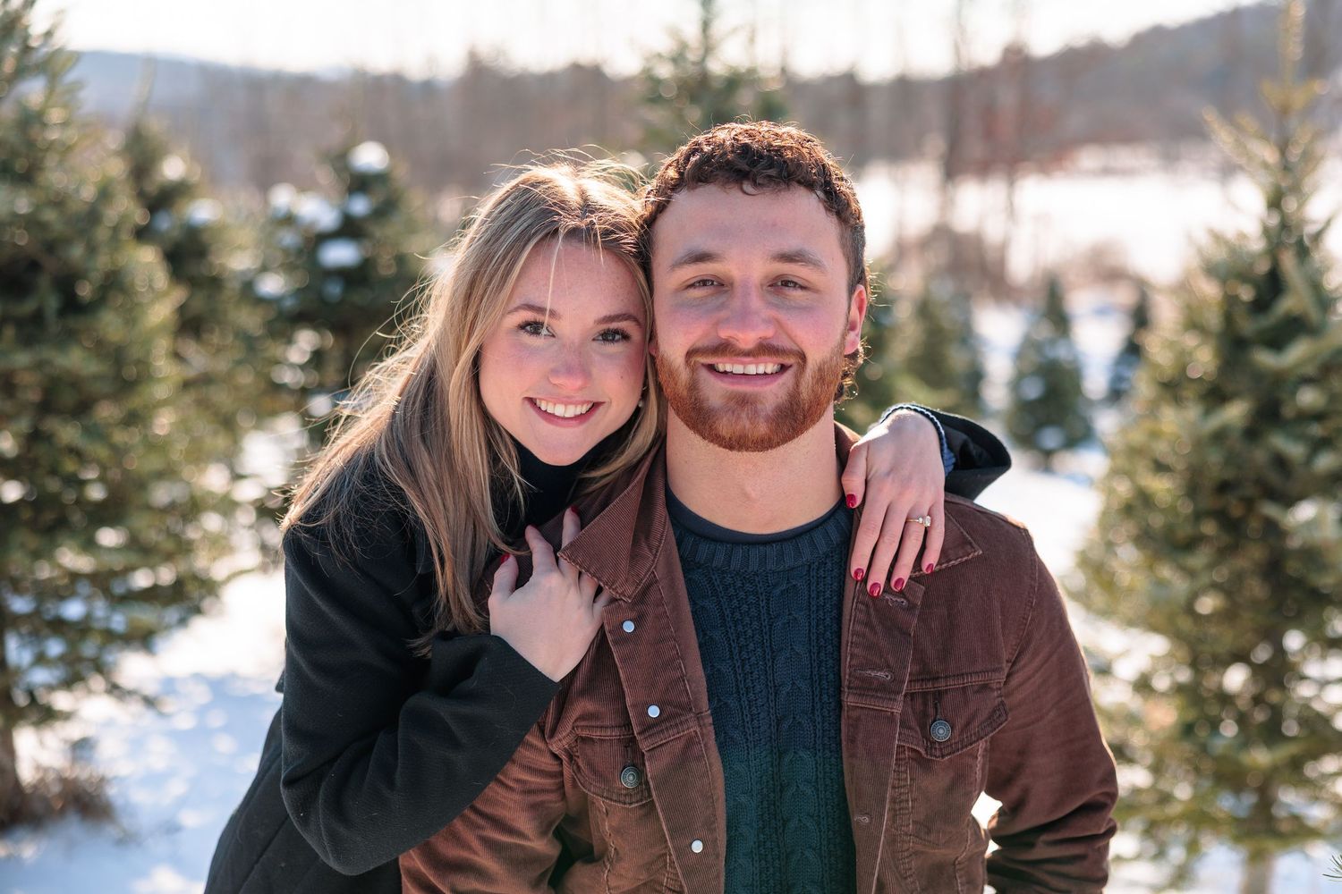 Smiling couple posing together at a snowy Christmas tree farm on a sunny winter day.