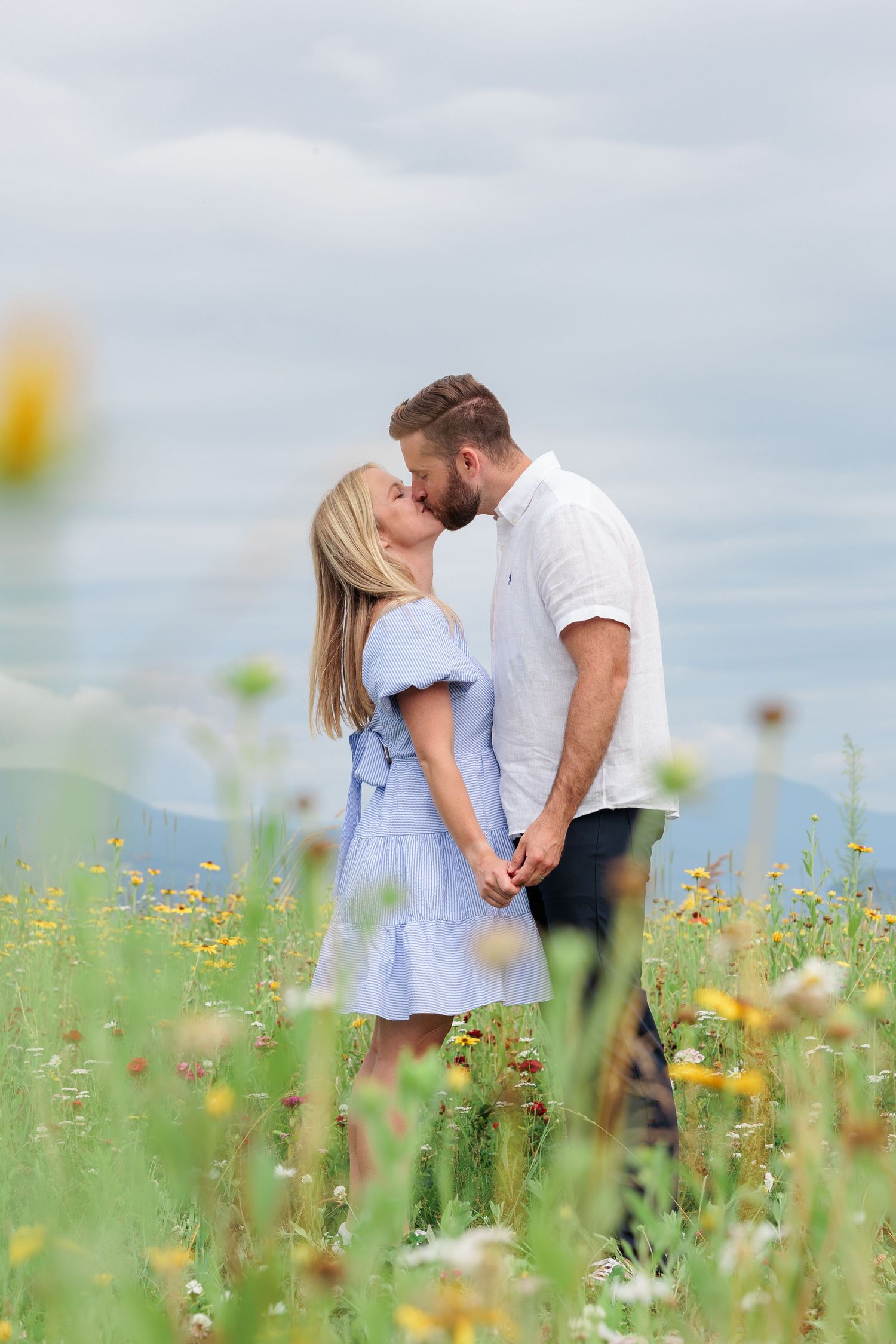 Couple sharing a romantic kiss in a wildflower field with mountains and cloudy sky in background.