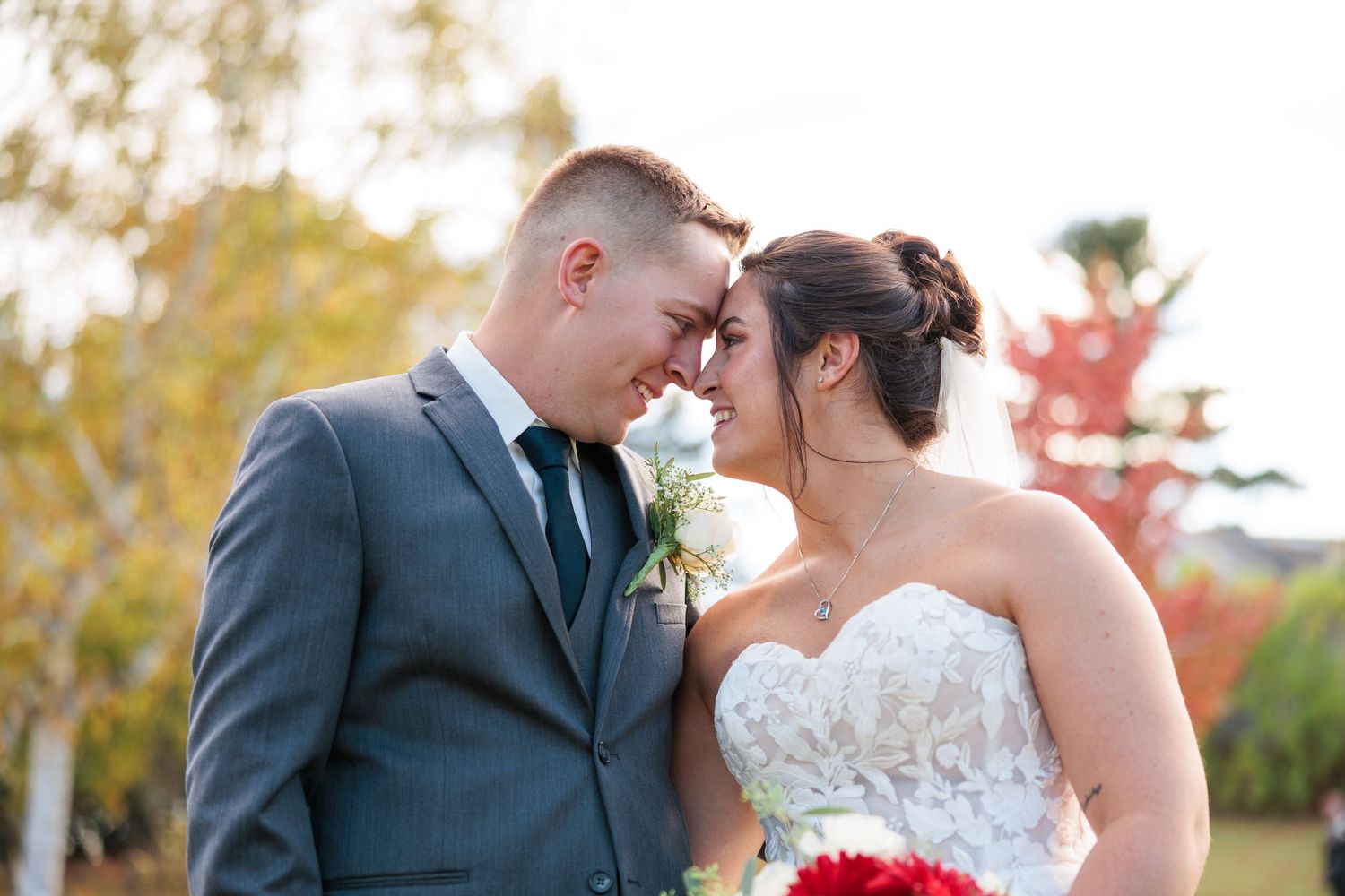 Bride and groom touch foreheads romantically outdoors surrounded by colorful red and yellow autumn foliage.