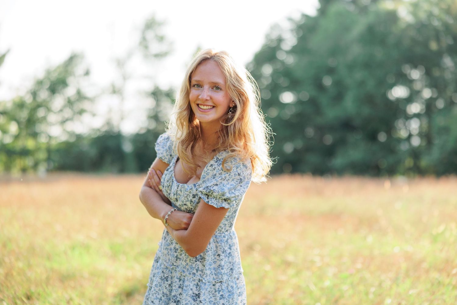 Smiling blonde woman in floral dress standing in golden field with arms crossed on sunny day.