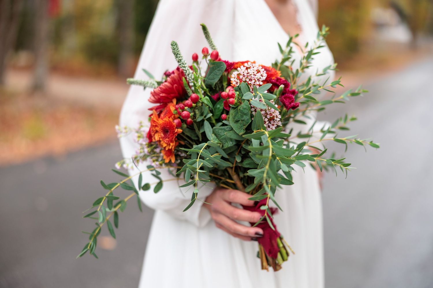 Bride holding a vibrant red and green wedding bouquet with roses, berries, and eucalyptus outdoors.