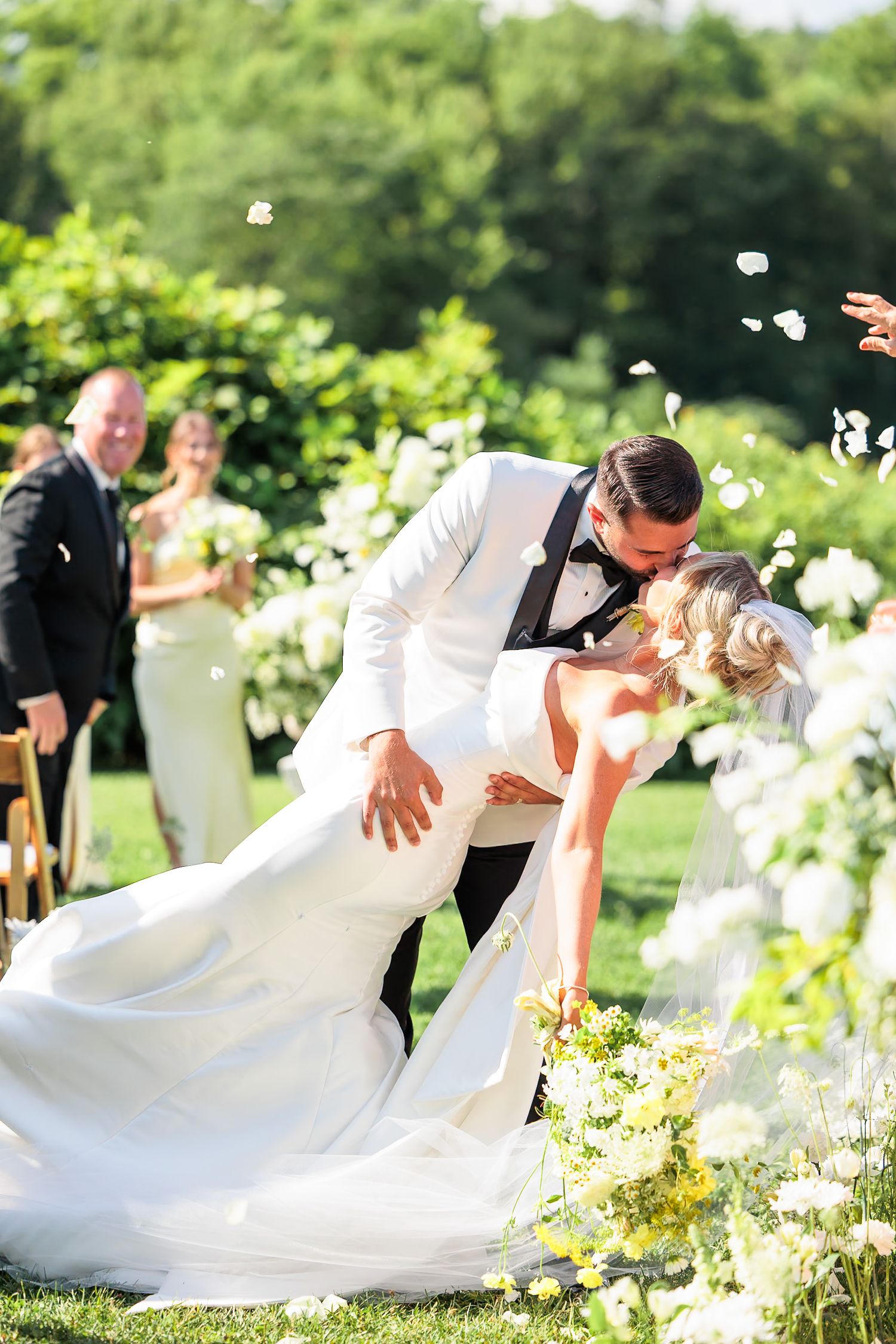 Groom dips bride for a kiss as rose petals fall during outdoor garden wedding ceremony.