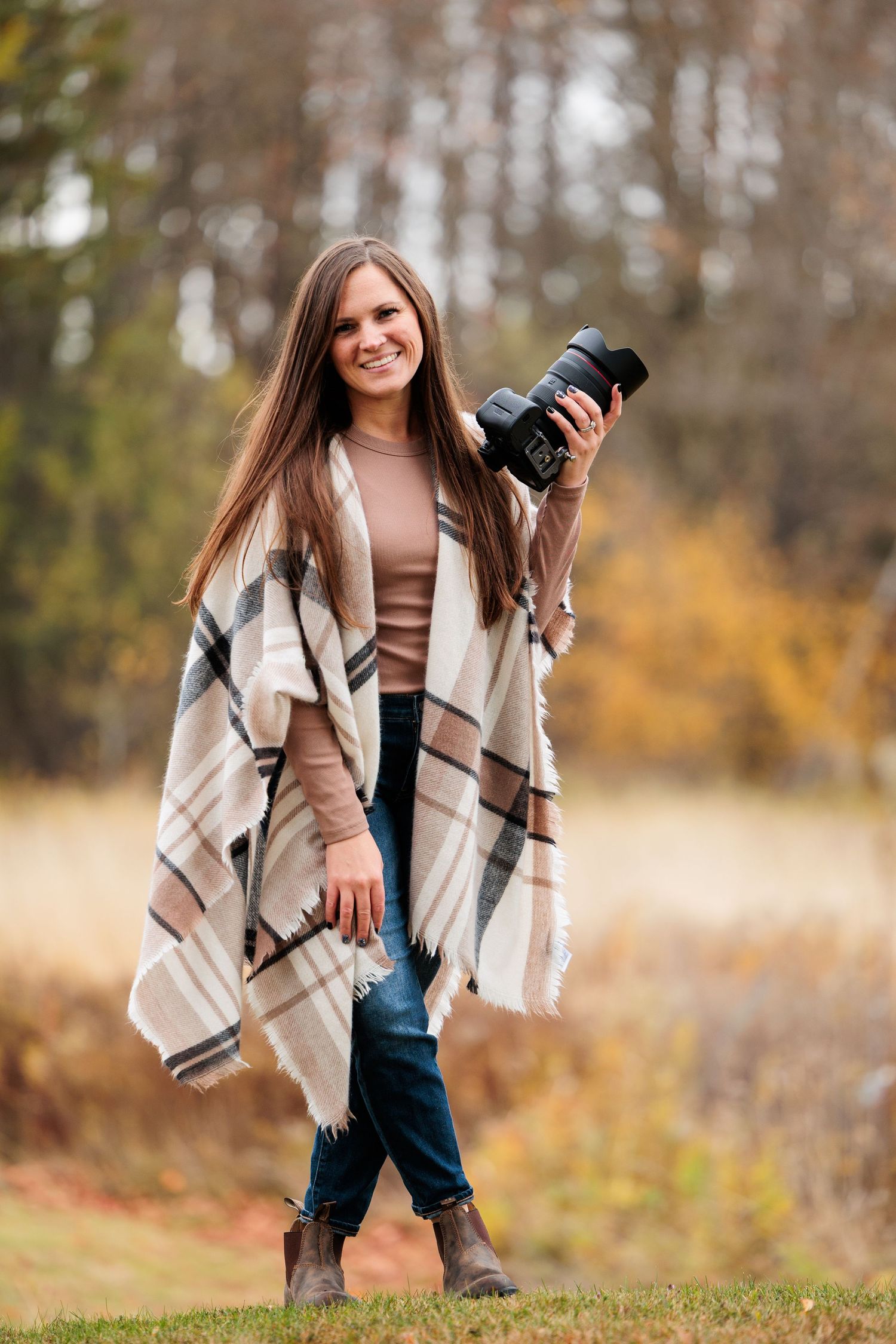 Smiling female wedding photographer poses outdoors in plaid wrap holding professional camera in fall.