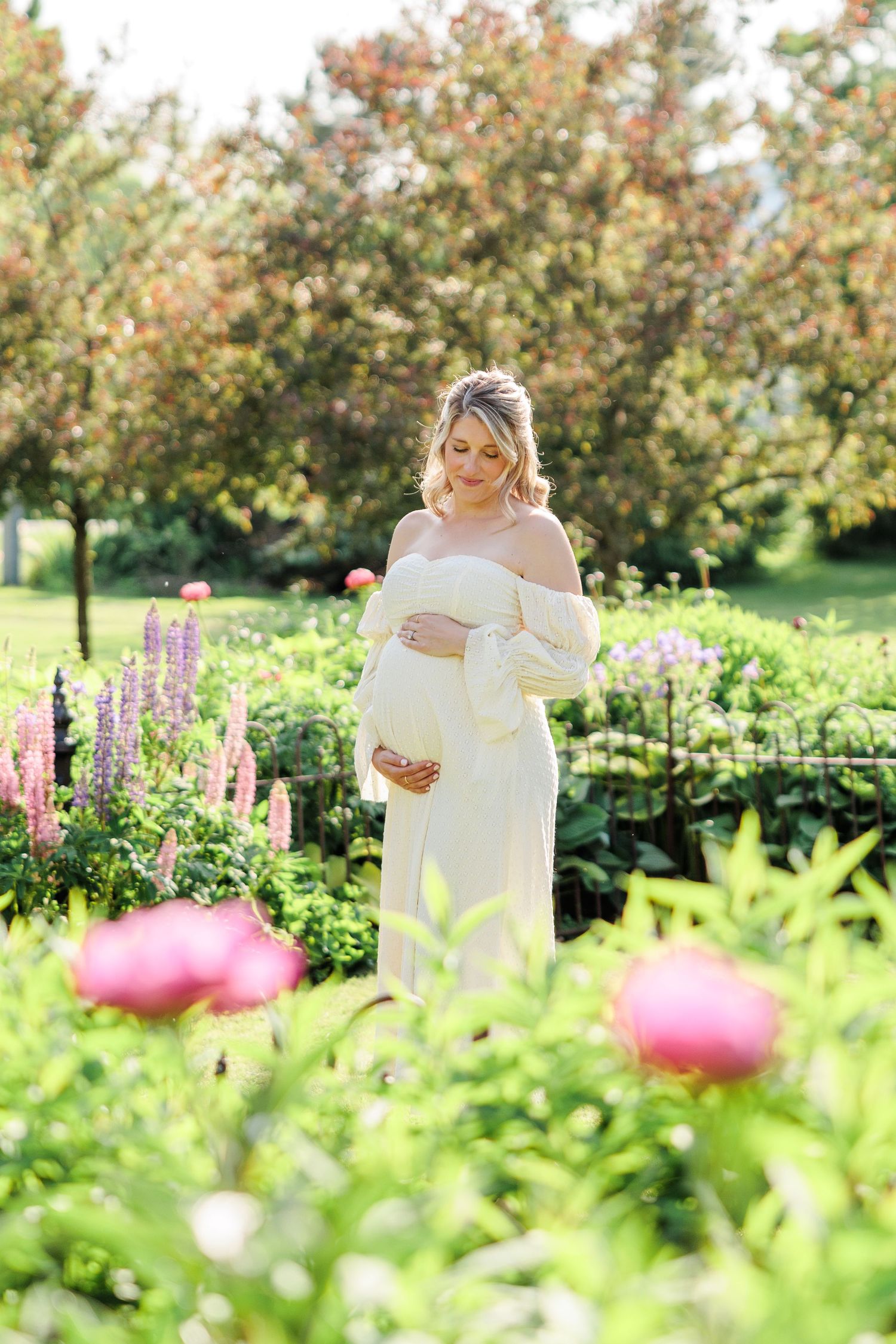Pregnant woman in a white off-shoulder gown cradling her belly in a lush garden with pink flowers.