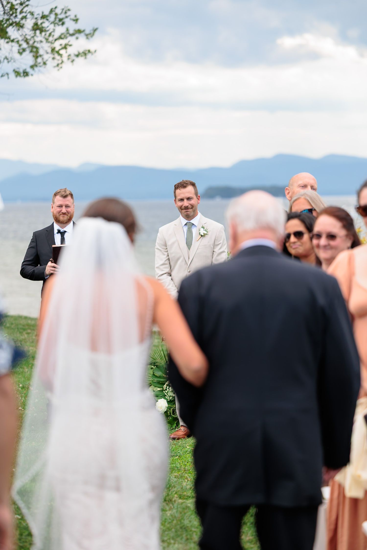 Groom in a beige suit smiling as he watches his bride walk down the aisle at an outdoor lakeside ceremony.