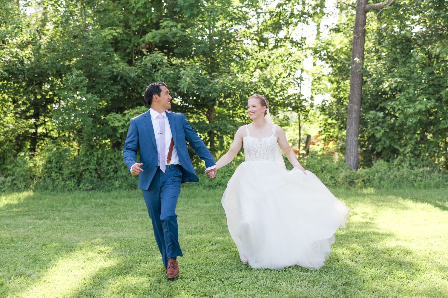Bride in flowing white gown and groom in navy suit holding hands running through lush green outdoor setting.