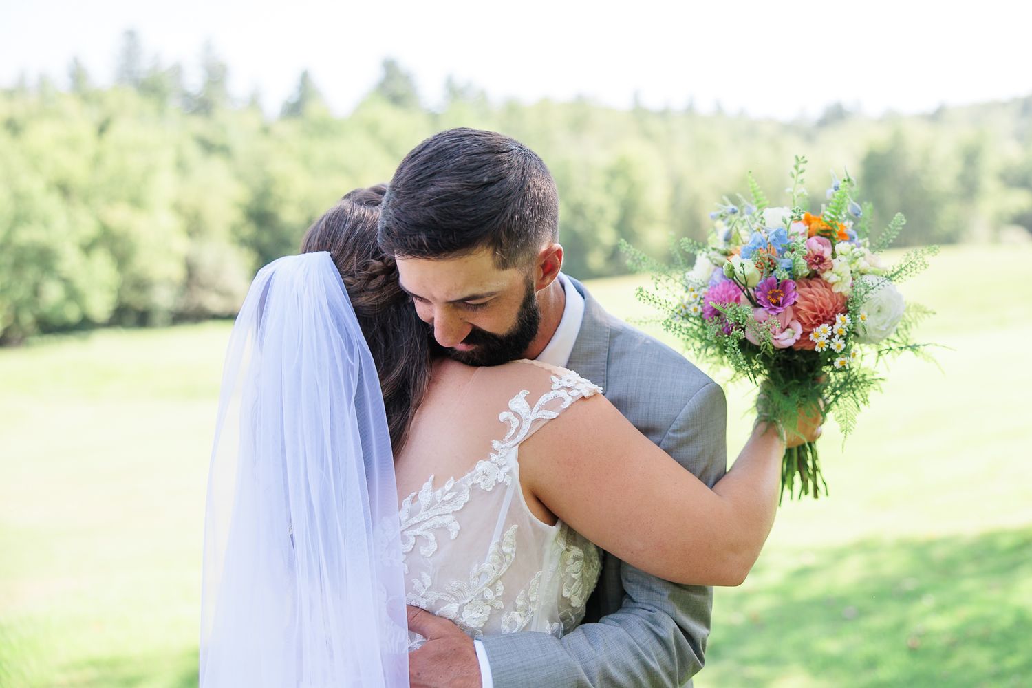 Groom embraces bride tenderly holding colorful bouquet on lush green lawn.