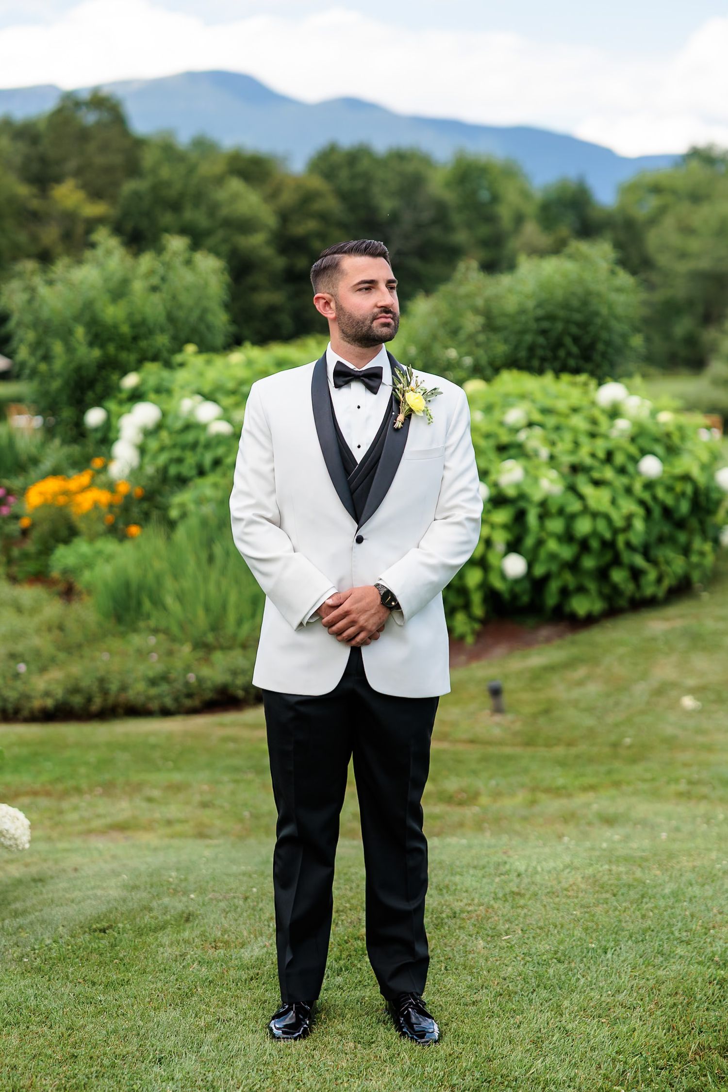 Handsome groom in white tuxedo jacket with boutonniere standing confidently in lush garden with mountain backdrop.