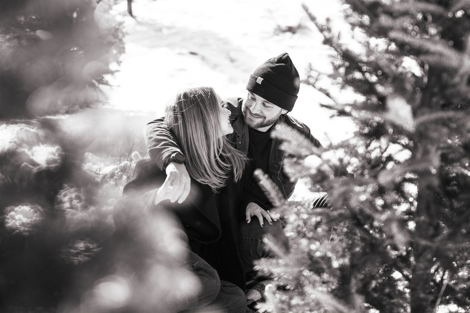 Black and white photo of a couple playfully hiding among snow-dusted Christmas trees.