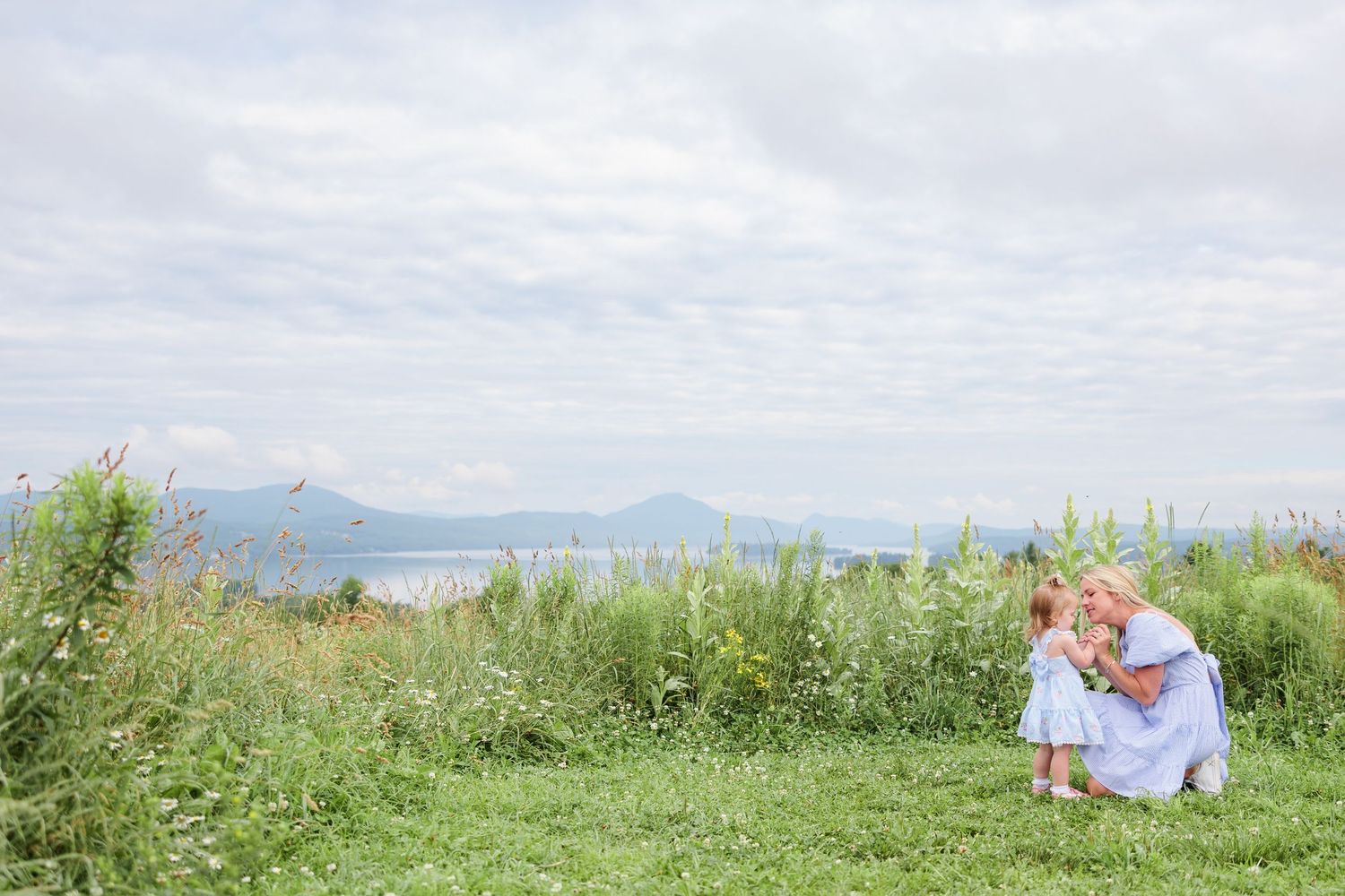Mother and young daughter sharing a tender moment in a green meadow with scenic lake backdrop.