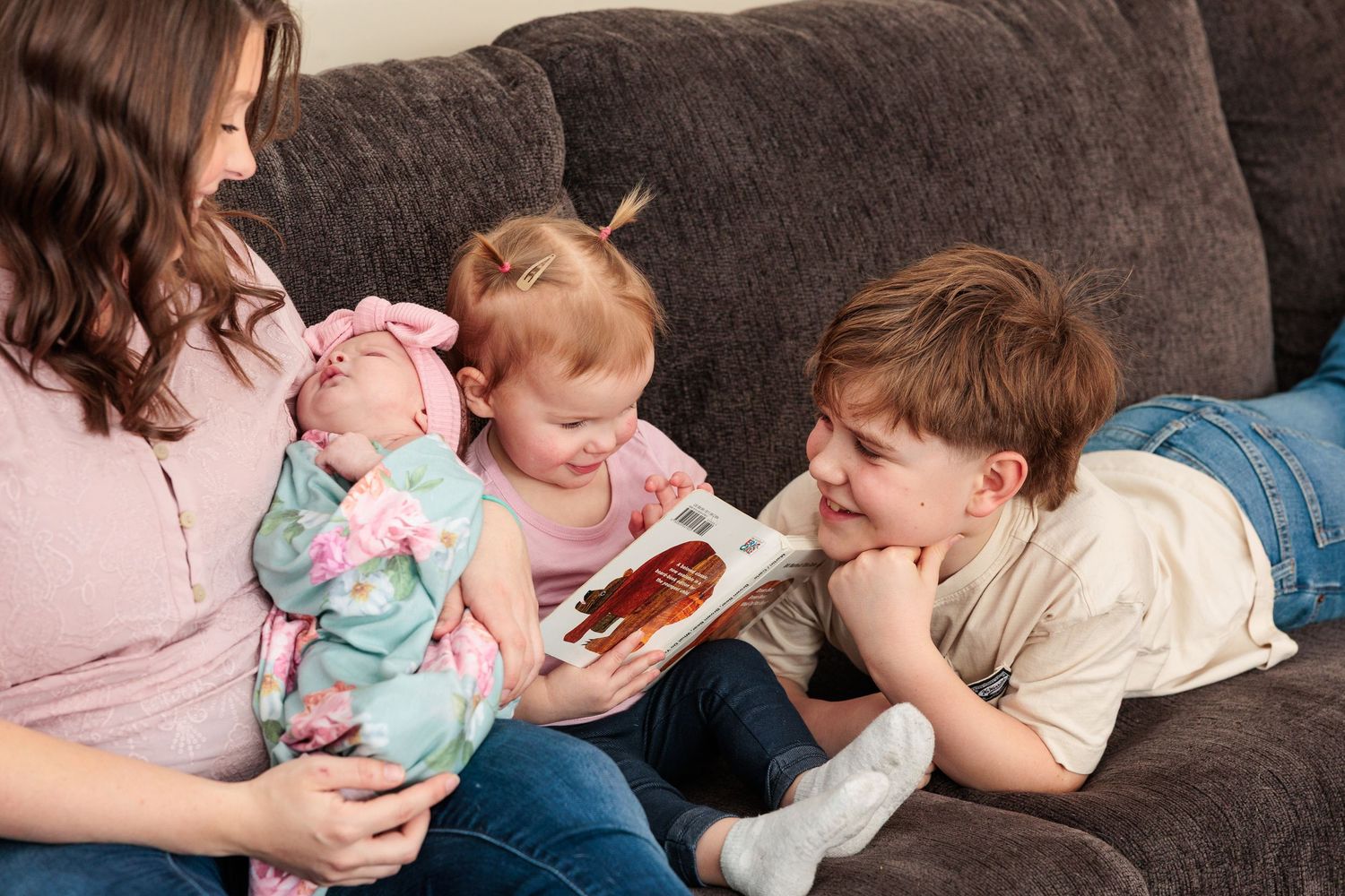 Mother holds newborn baby on couch while toddler daughter and young son read a picture book.