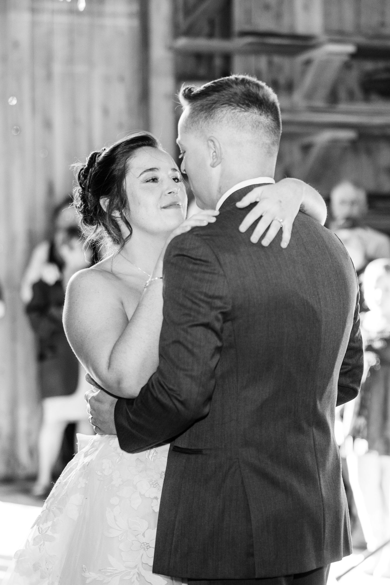 Black and white photo of bride smiling joyfully while dancing with groom at barn reception.