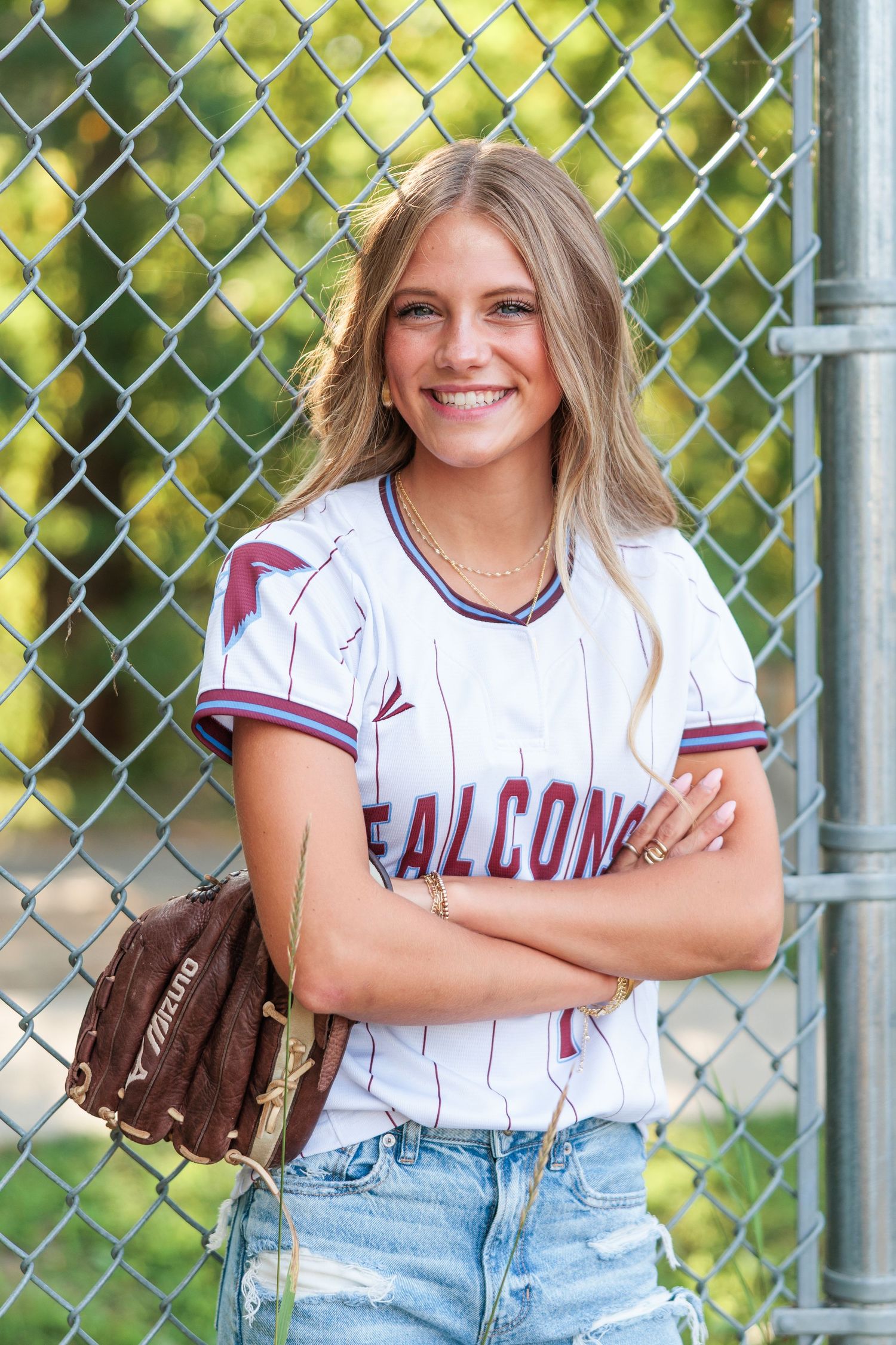 Smiling young woman in Falcons softball jersey with mitt leaning against chain-link fence.