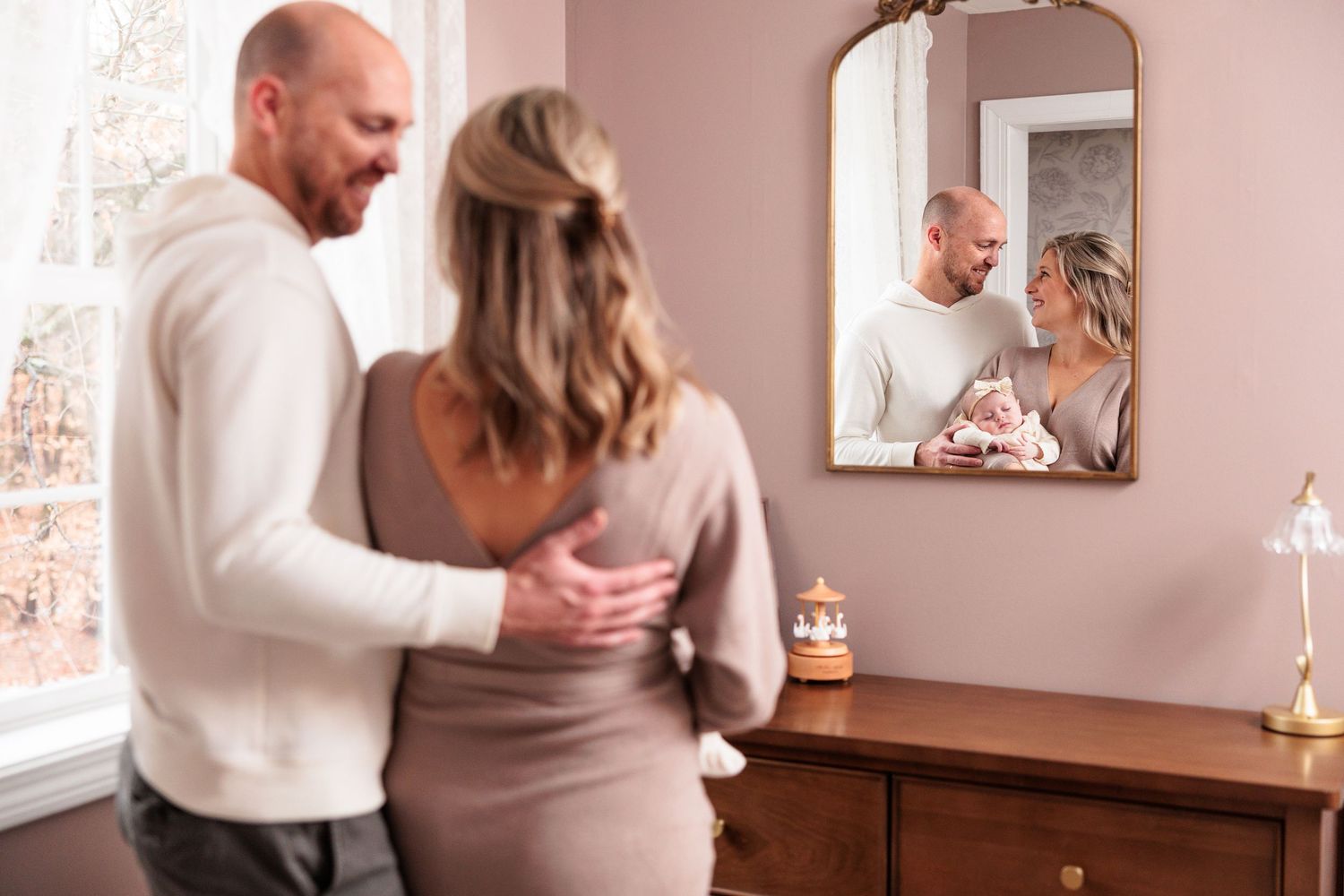Parents gazing at their reflection in vintage mirror while standing beside dresser in nursery.