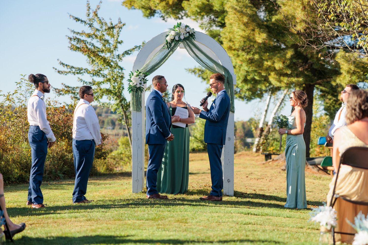 Outdoor wedding ceremony under white arch with guests gathered on grassy hillside.
