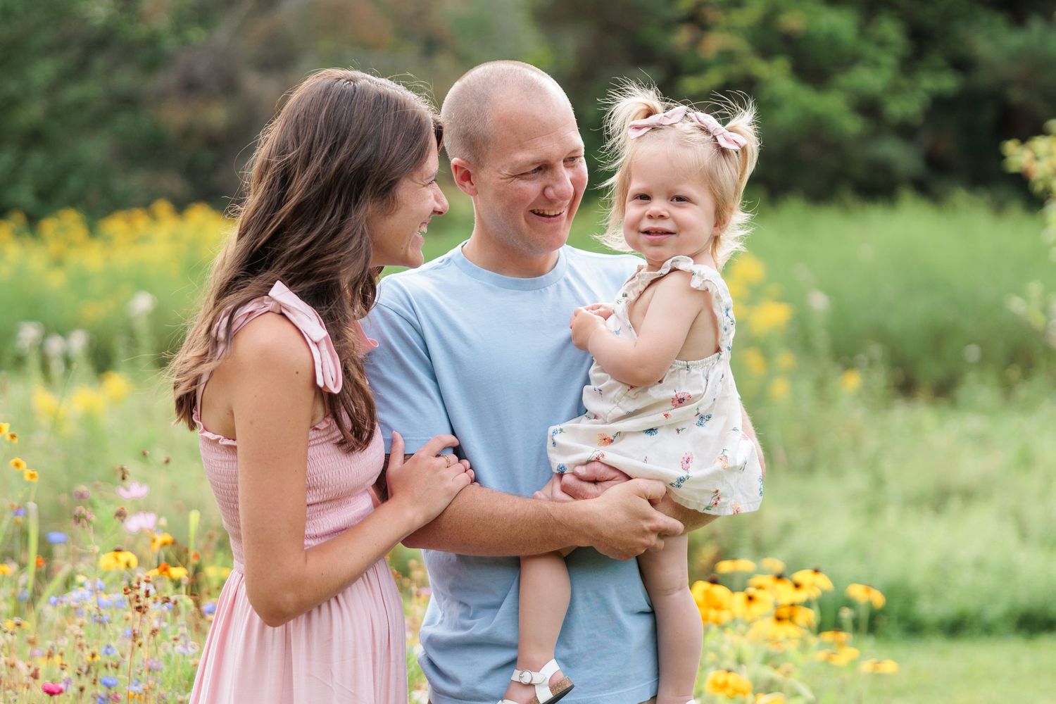 Smiling parents hold their toddler daughter in a sunny field of yellow wildflowers.