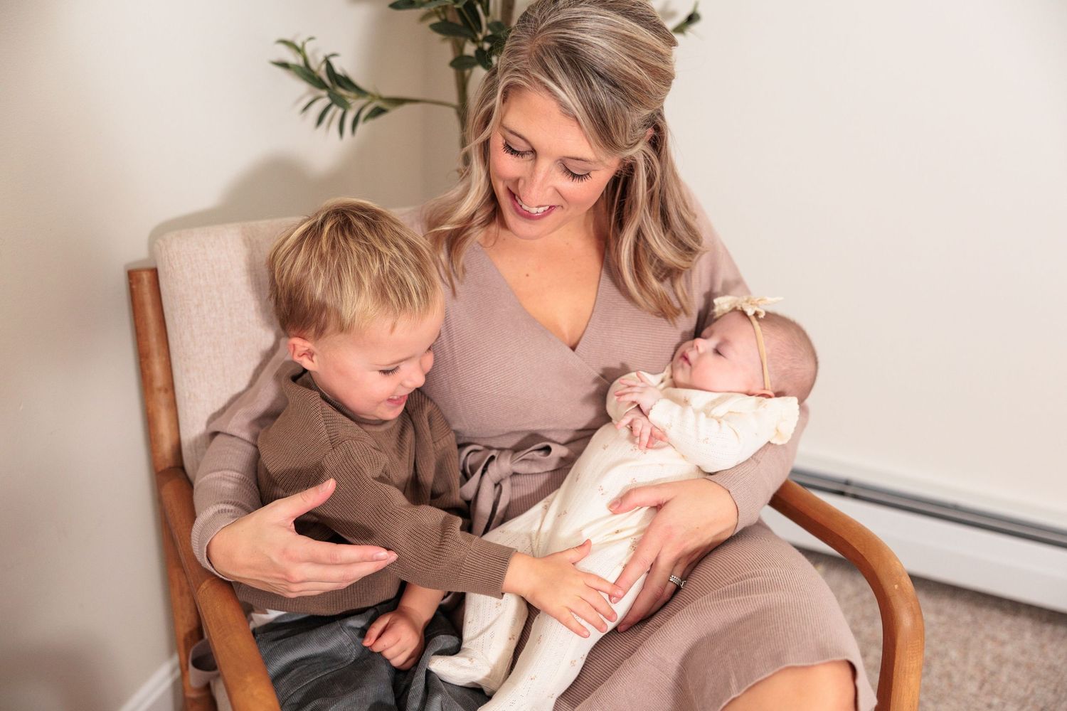 Smiling blonde mother in mauve dress sitting in chair holding newborn while young son leans in closely.