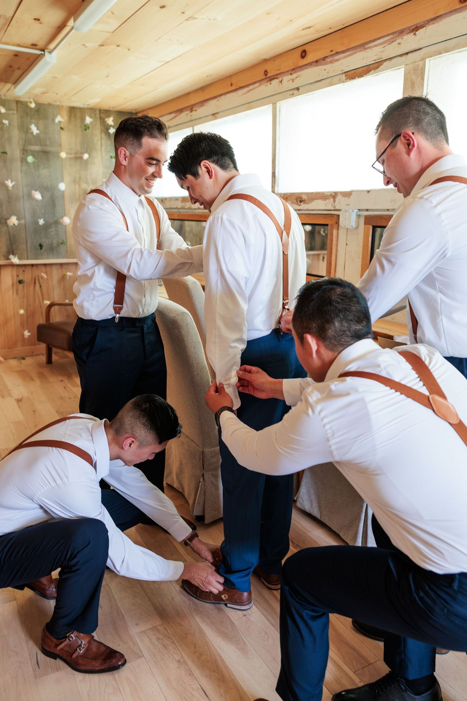 Groomsmen in white shirts and suspenders help groom get dressed in a rustic wooden venue.