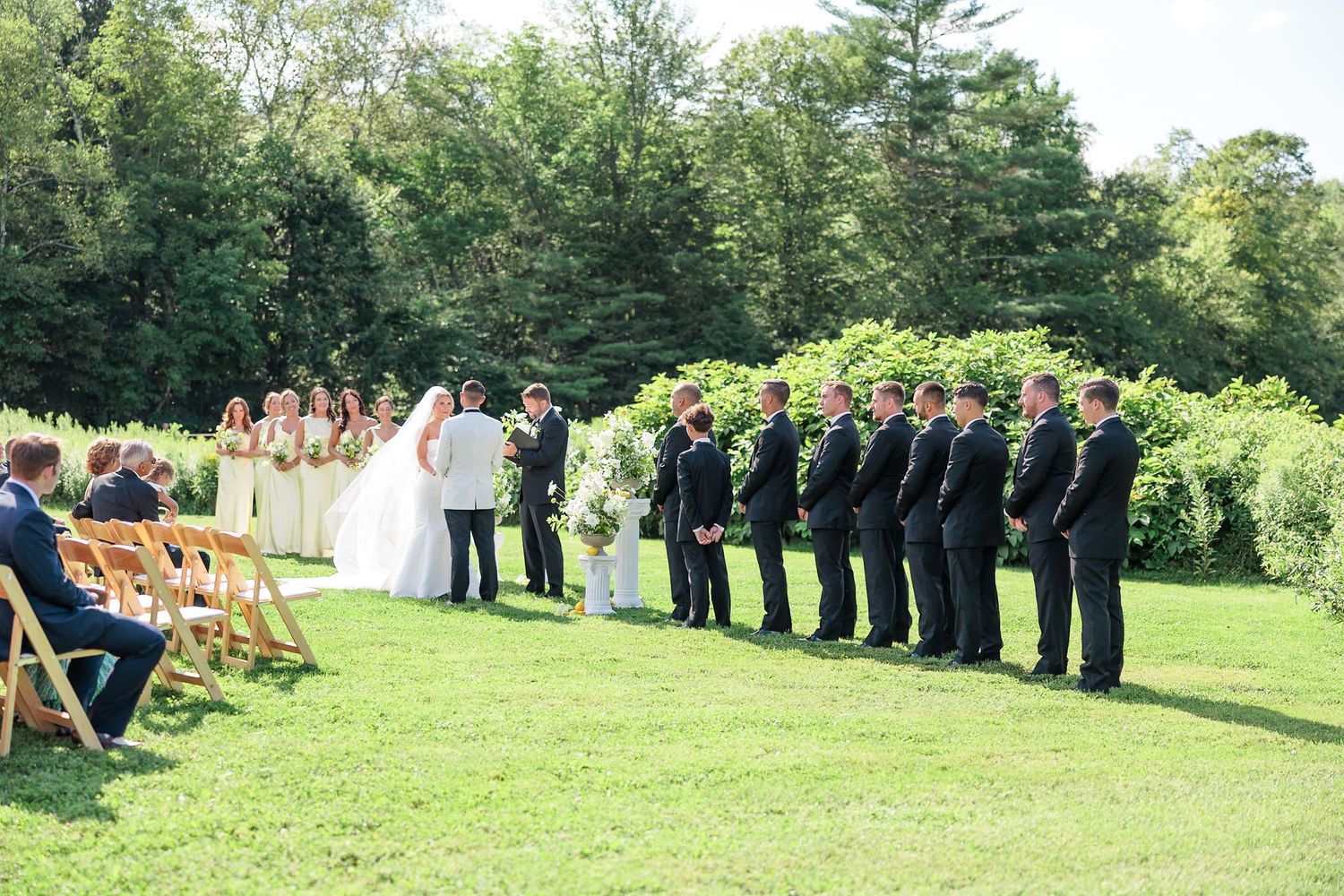 Outdoor wedding ceremony on green lawn with groomsmen in black suits and bridesmaids in yellow gowns.