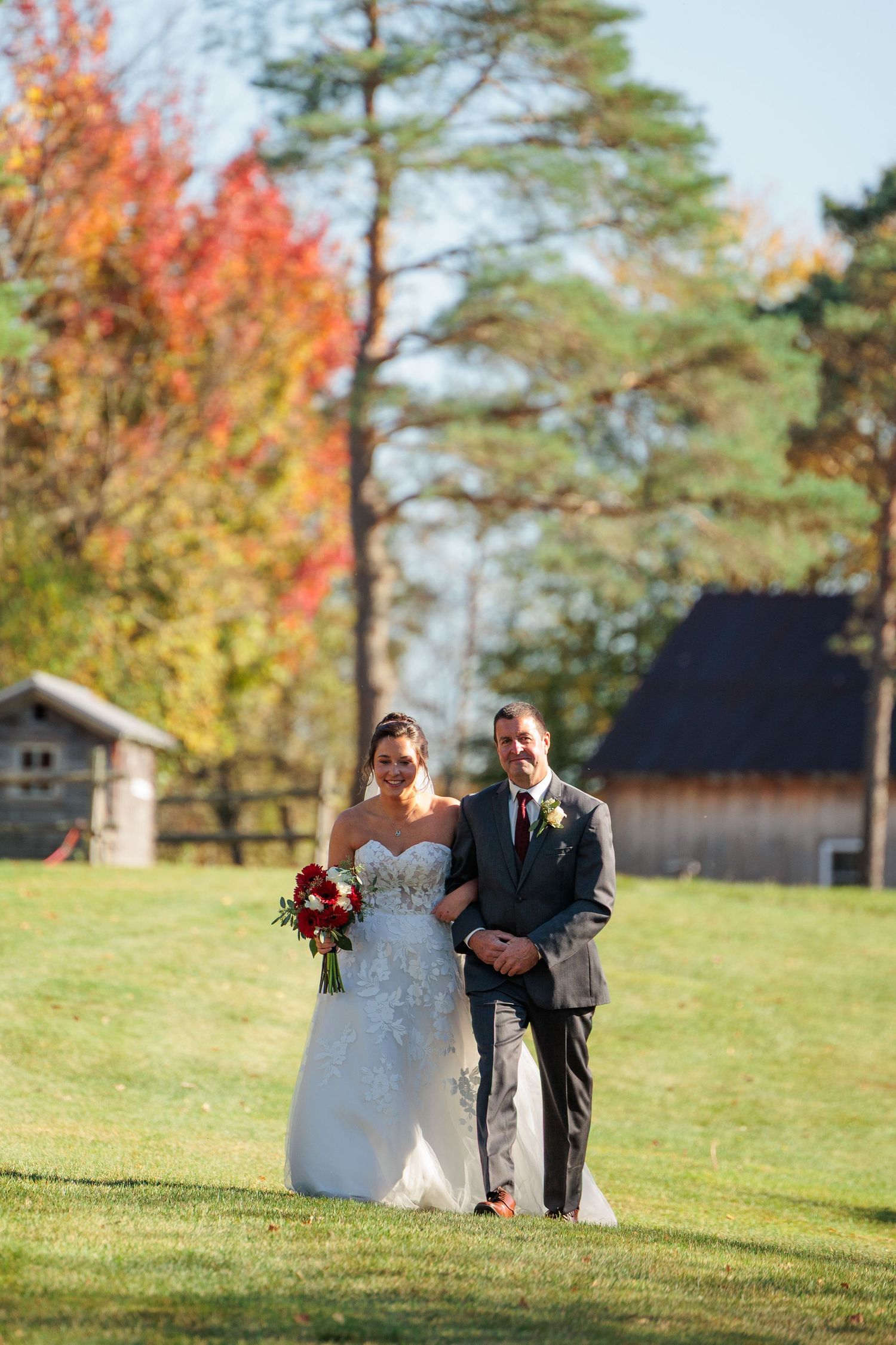 Bride and groom walk together on green lawn with vibrant autumn foliage backdrop at outdoor fall wedding.