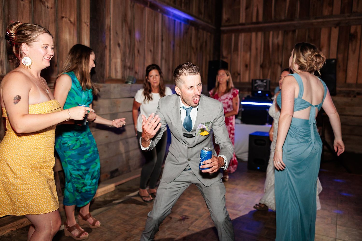 Groom in gray suit dances energetically with guests on wooden dance floor at rustic barn wedding.