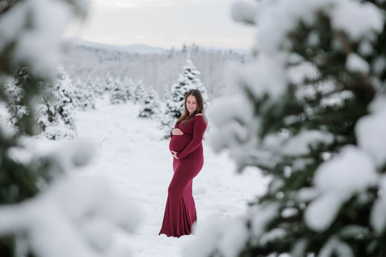 A pregnant woman in a red gown smiles softly while holding her bump in a snowy pine tree setting.