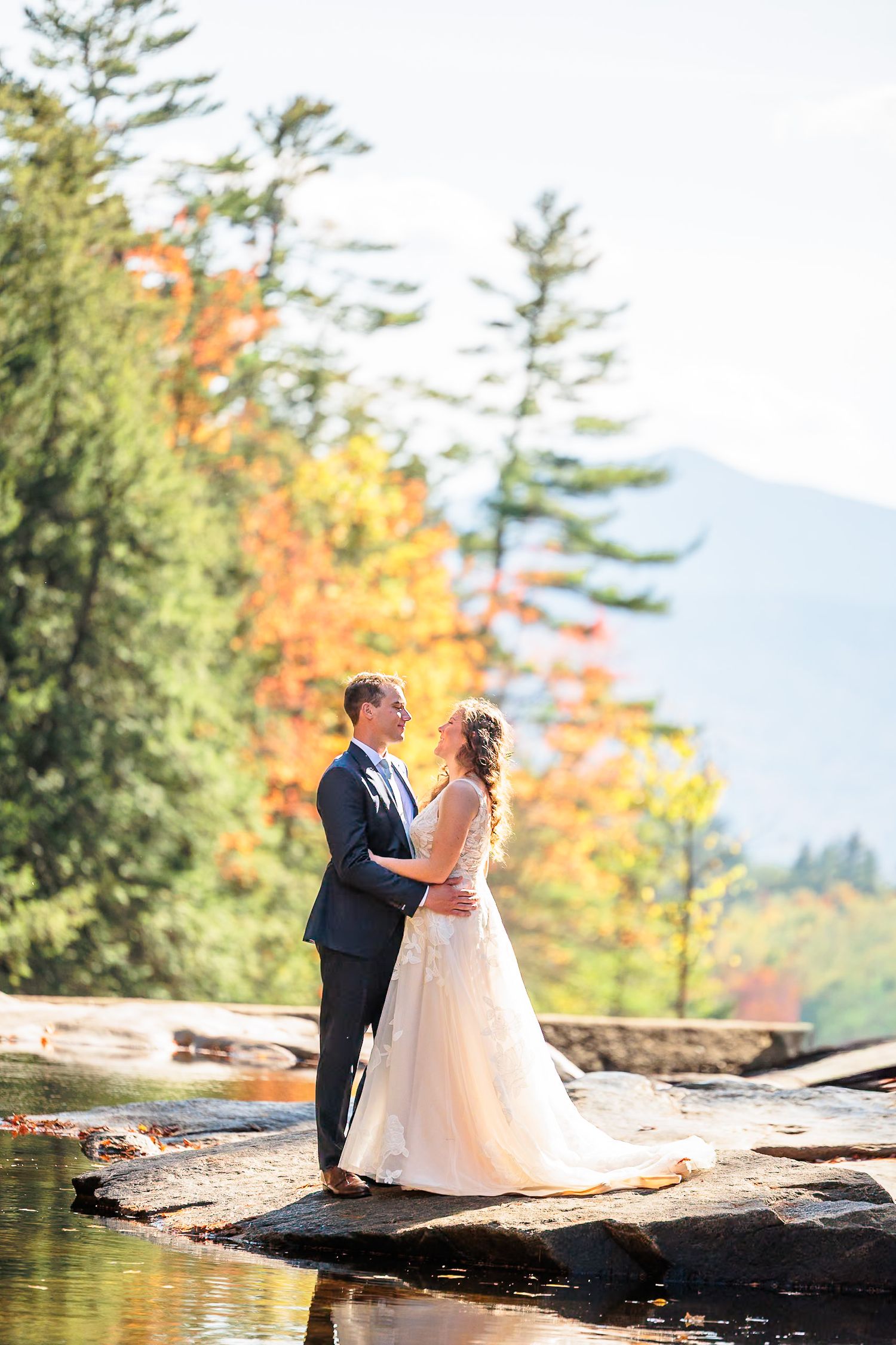 Couple shares tender moment on rocky riverbank surrounded by vibrant autumn foliage and mountains.