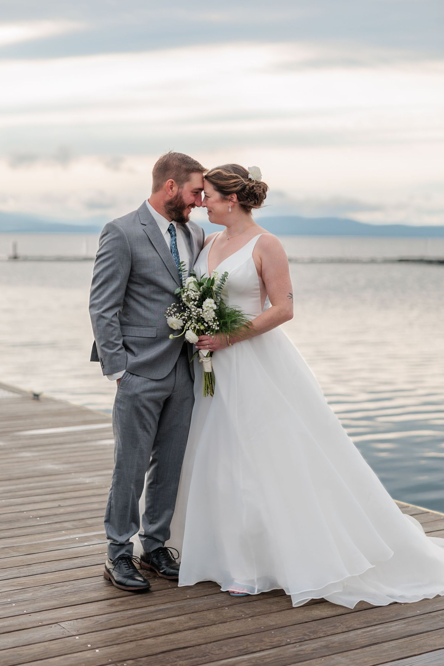 Bride and groom share a romantic moment on a wooden dock by the water, she holds a white bouquet in a flowing gown.