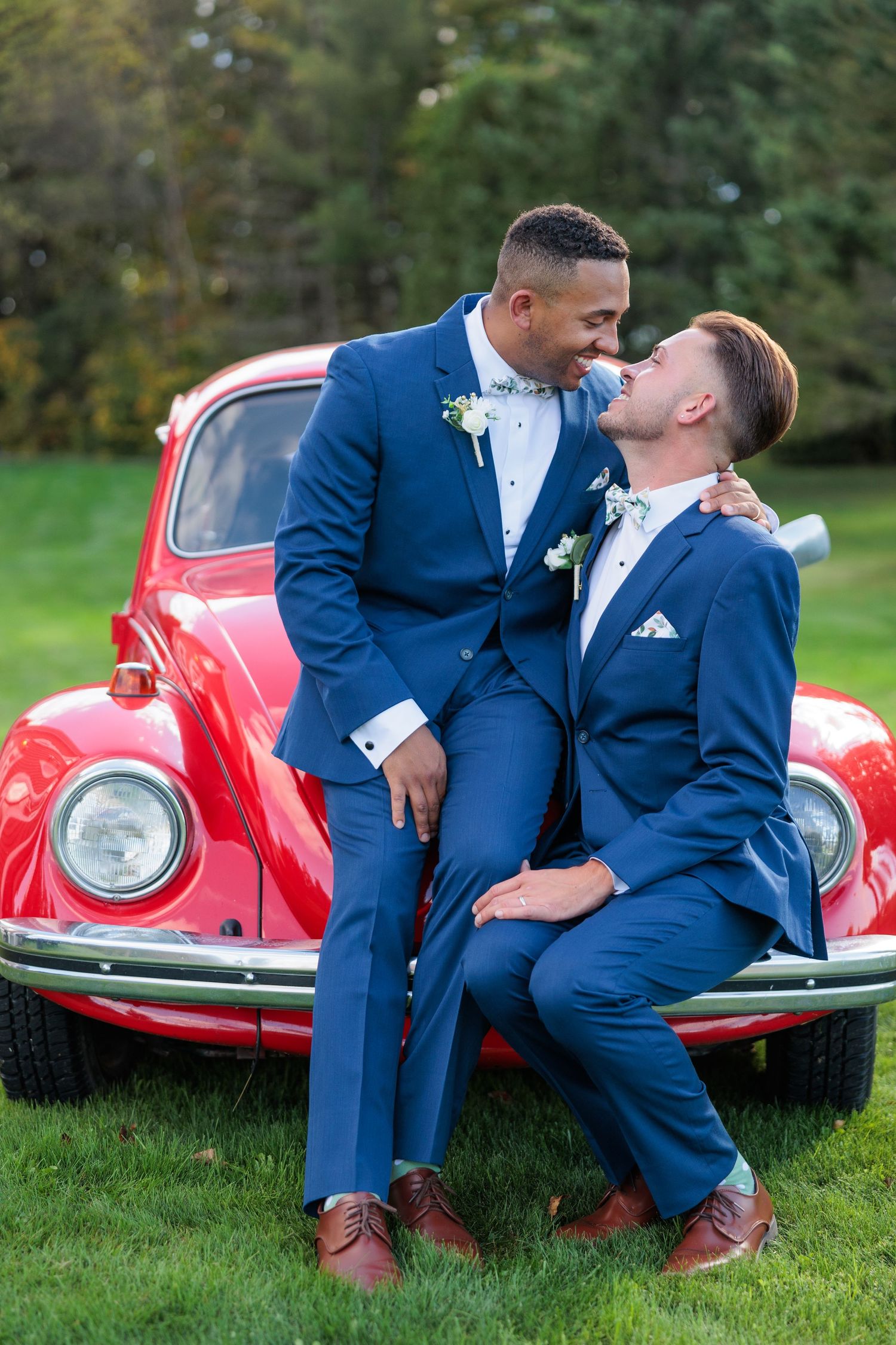 Wedding couple in blue suits pose intimately against red classic Beetle on grass.