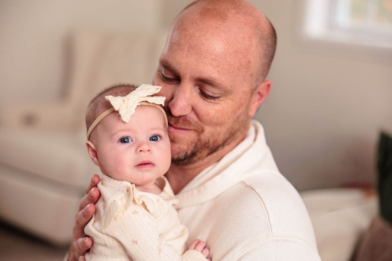 Sweet moment between parent and infant in white clothing against neutral background.