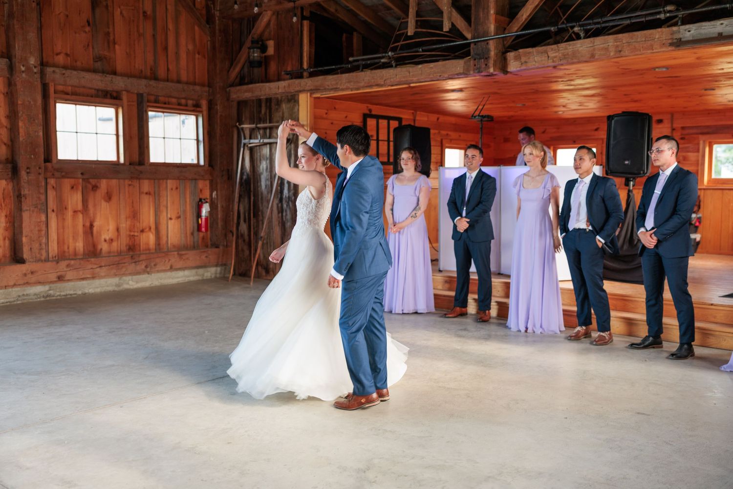 Bride and groom share first dance in rustic barn wedding venue while wedding party watches in formal attire.
